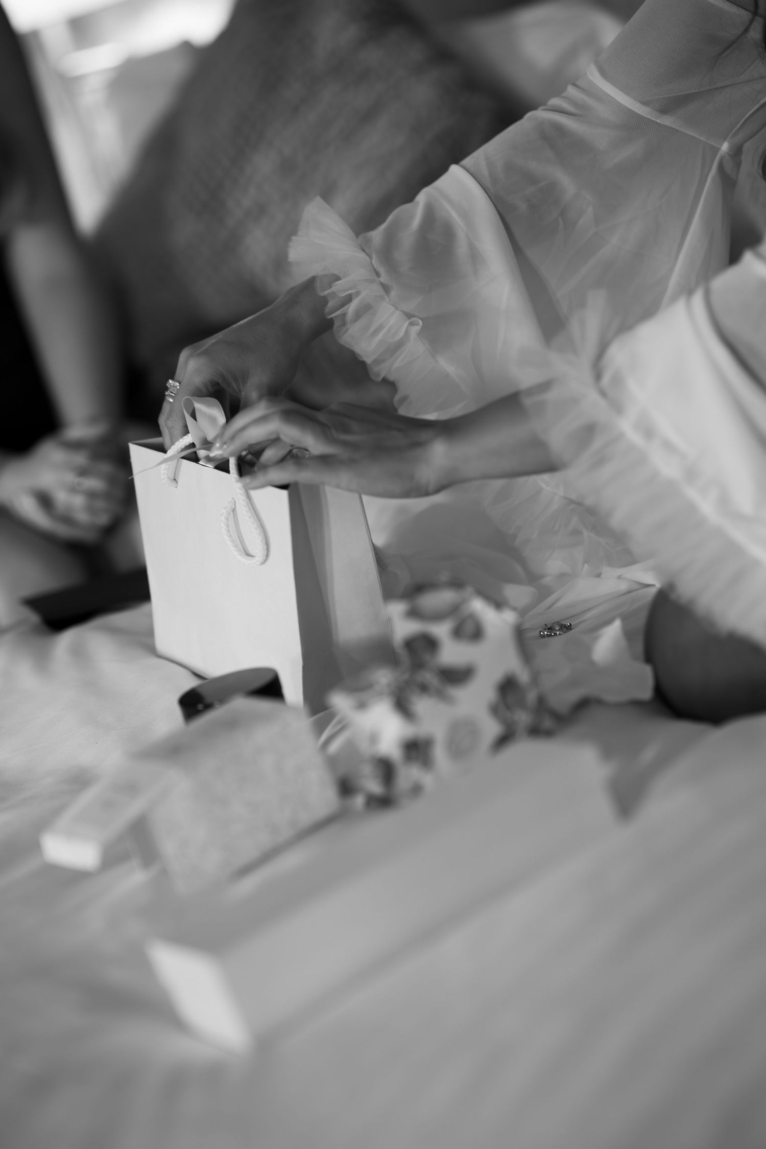 Black and white photo of a bride in white robe holding a small gift bag on a bed with wrapped presents.