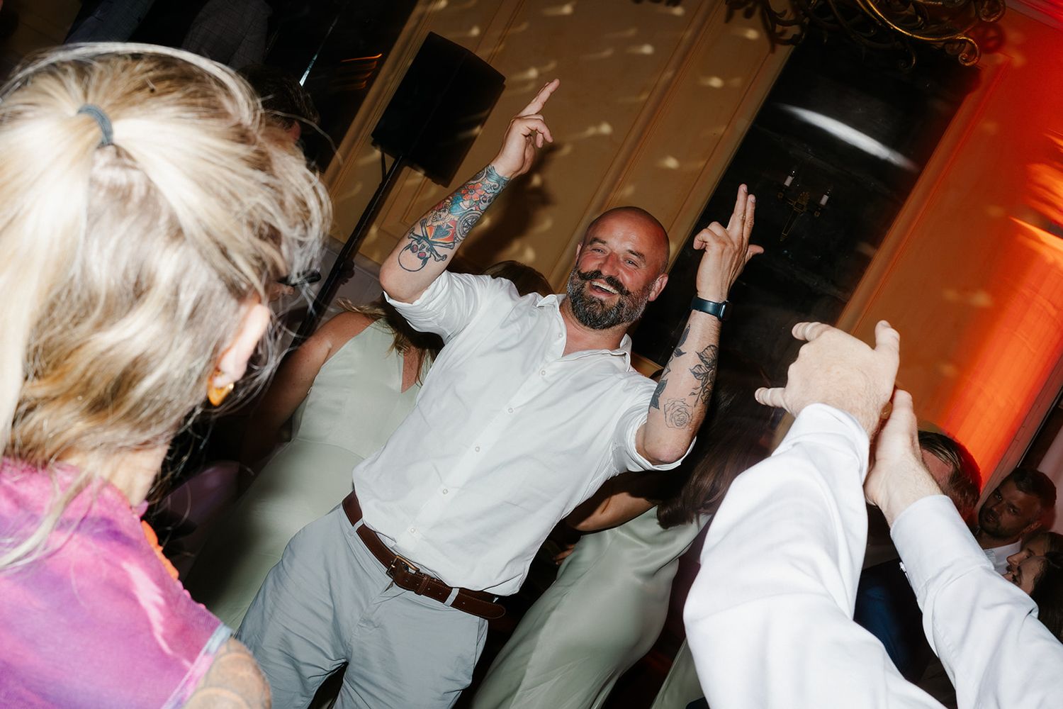 Tattooed man dancing joyfully at a wedding reception, raising hands with guests on the dance floor.