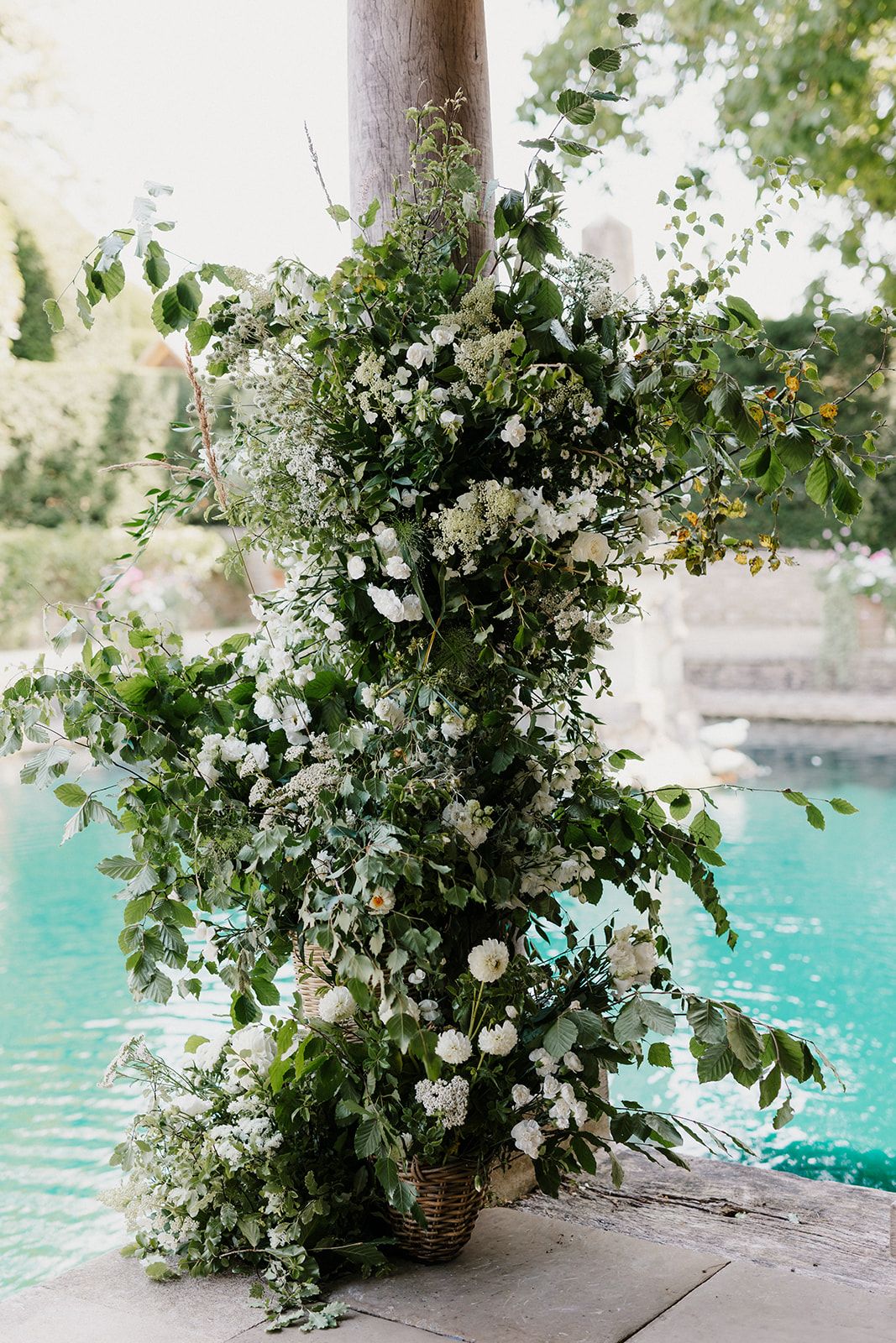 Lush white floral arrangement with greenery in a wicker basket beside a turquoise pool at an outdoor wedding.
