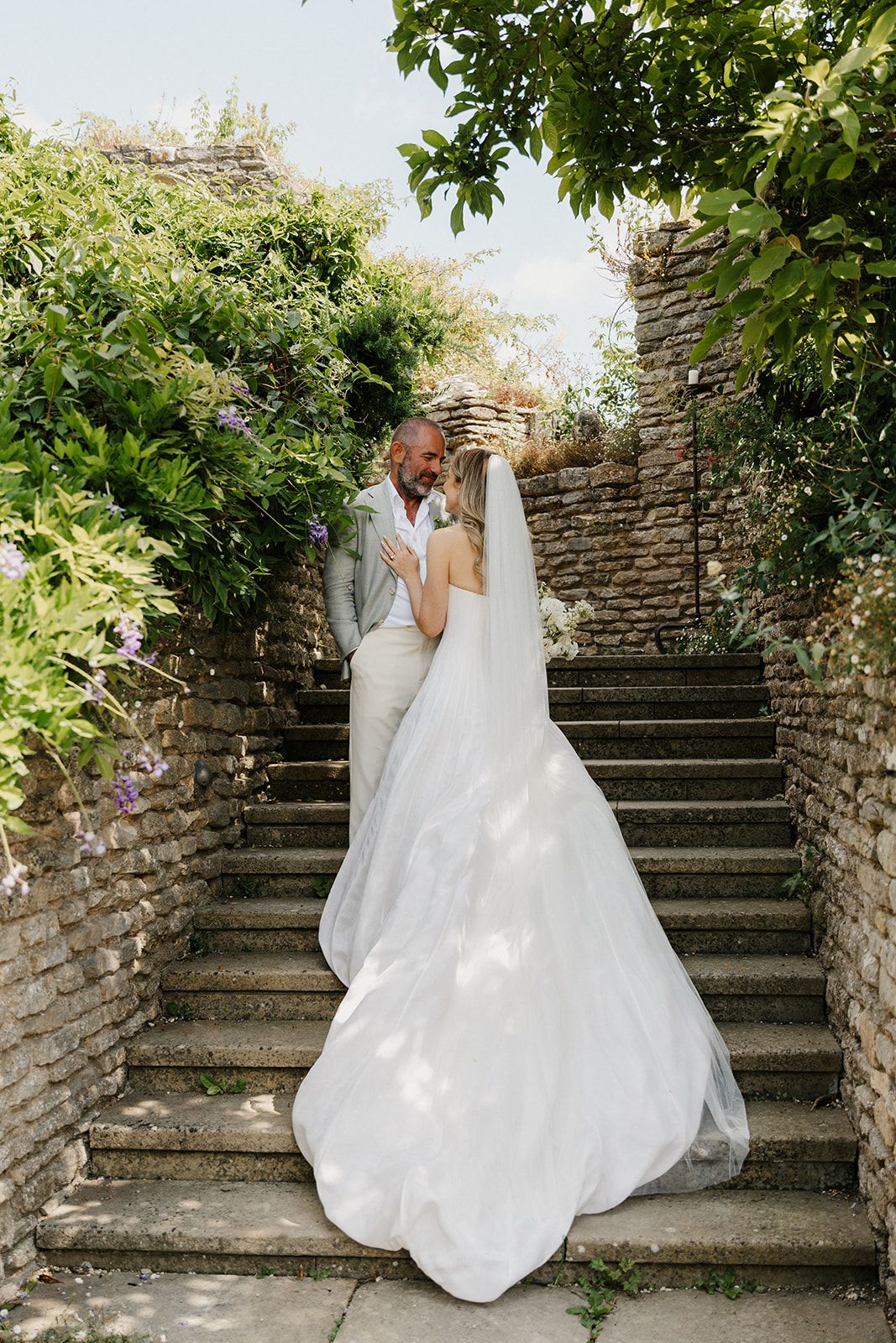Bride and groom share a romantic moment on stone steps surrounded by lush greenery, bride in flowing white gown with veil.