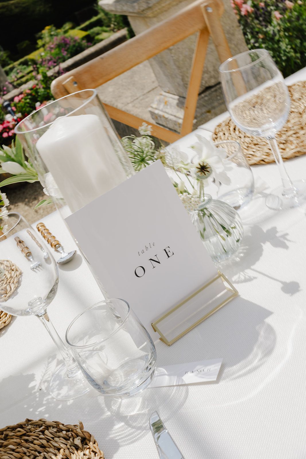 Elegant outdoor wedding table setting with white candle, floral arrangement, and Table One card on white linen.