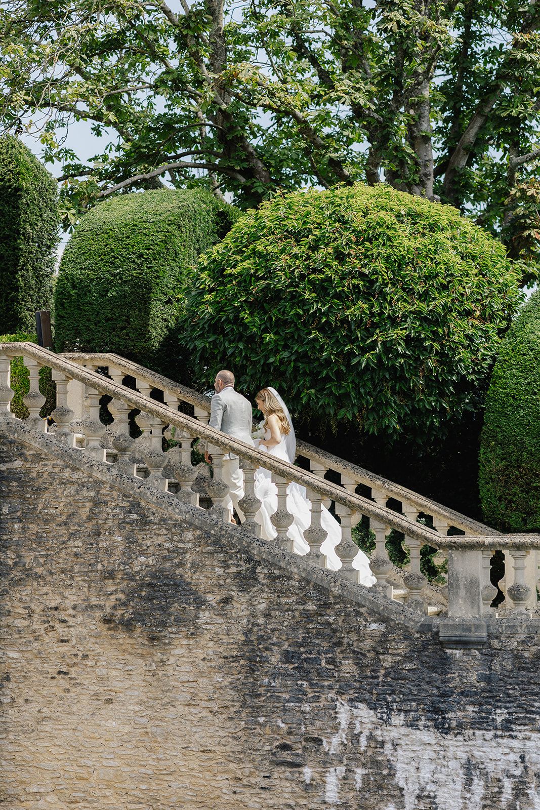 Bride and groom walking up stone balustrade staircase surrounded by lush manicured hedges in formal garden.