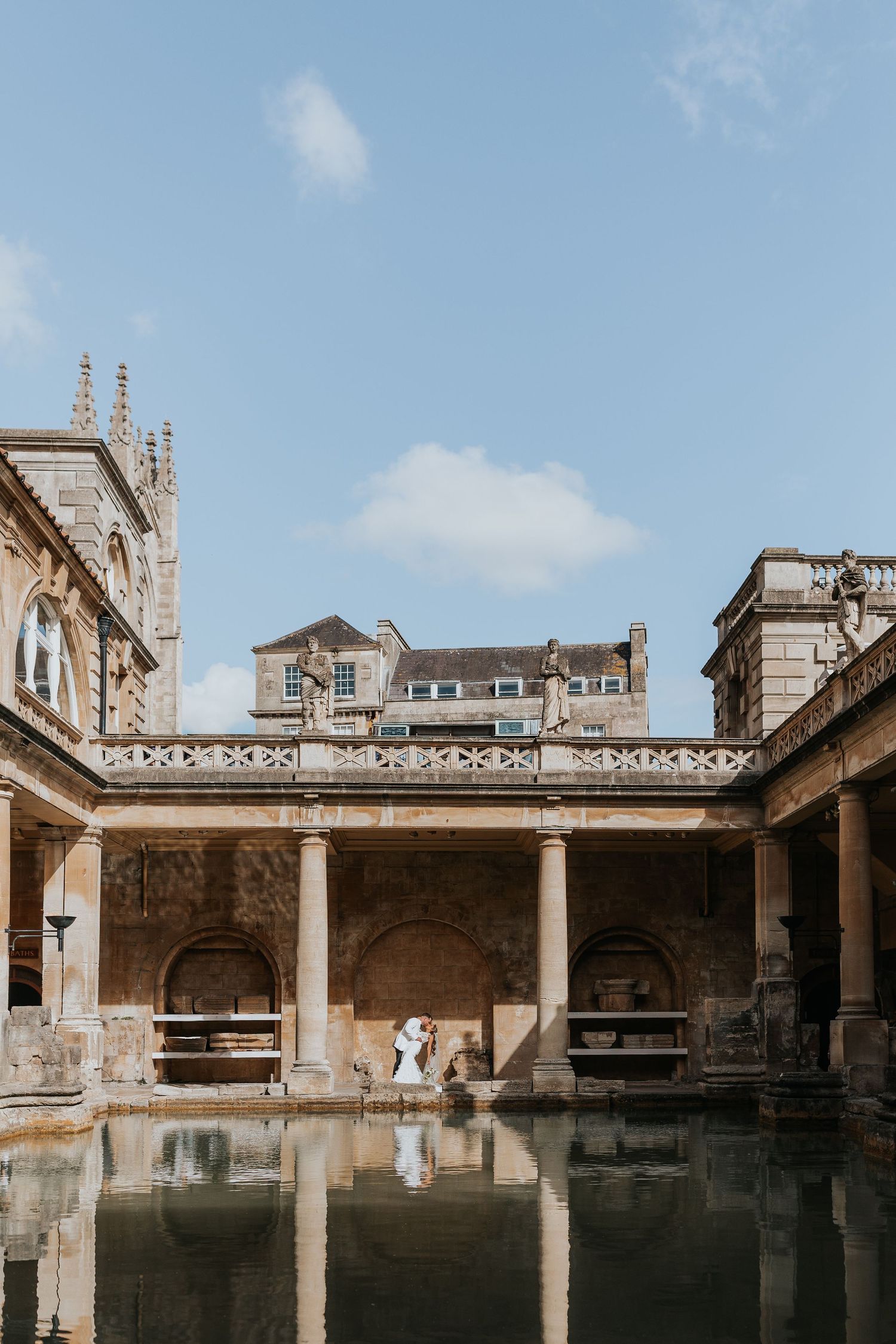 Couple sharing a romantic kiss at the historic Roman Baths in Bath, England, reflected in the ancient green waters.
