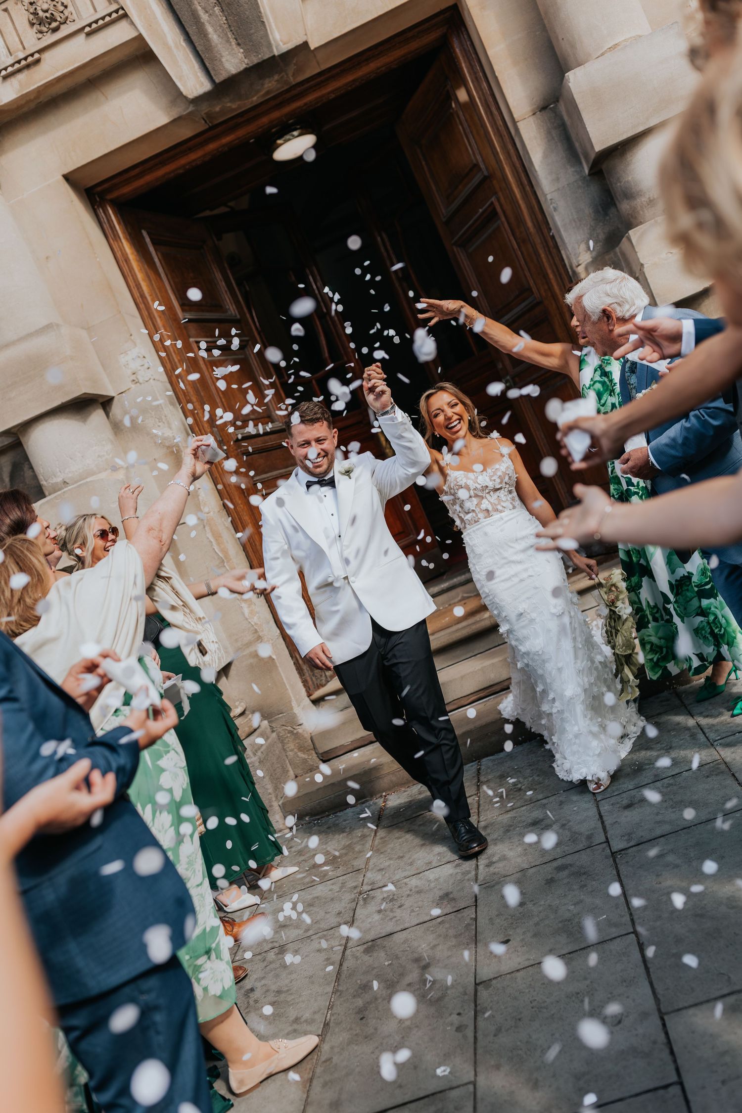 Bride and groom exit church doors as guests throw white confetti during joyful outdoor wedding ceremony celebration.