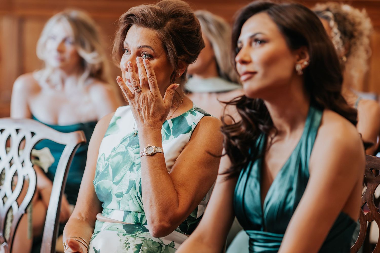 Emotional wedding guest wipes tears while seated next to bridesmaid in teal dress during ceremony.