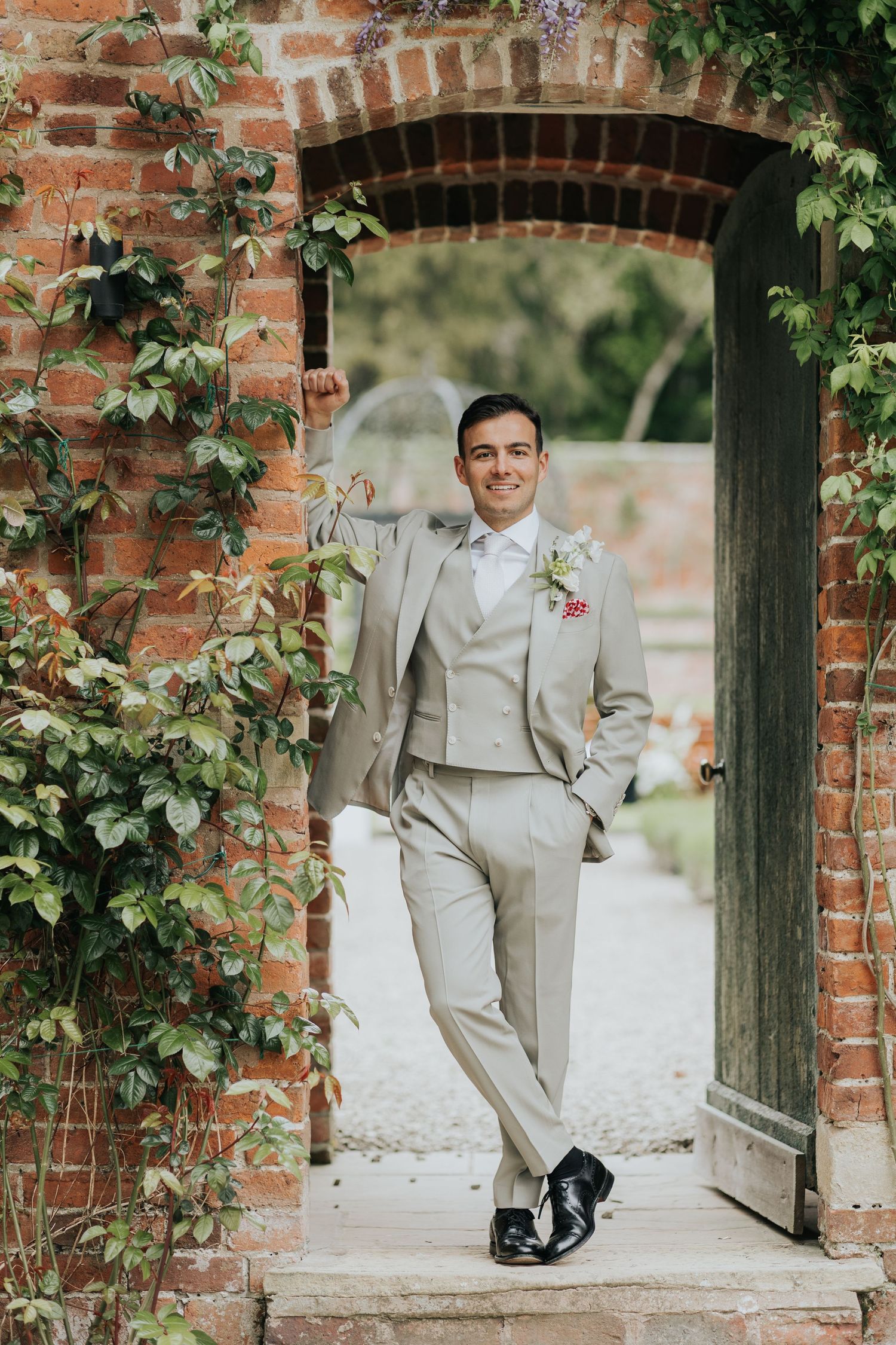 Smiling groom in light gray suit with boutonniere poses in ivy-covered brick archway doorway at wedding venue.