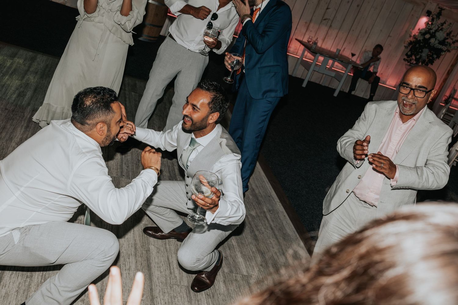 Men in formal white and navy suits socializing at a wedding reception, holding drinks on the dance floor.