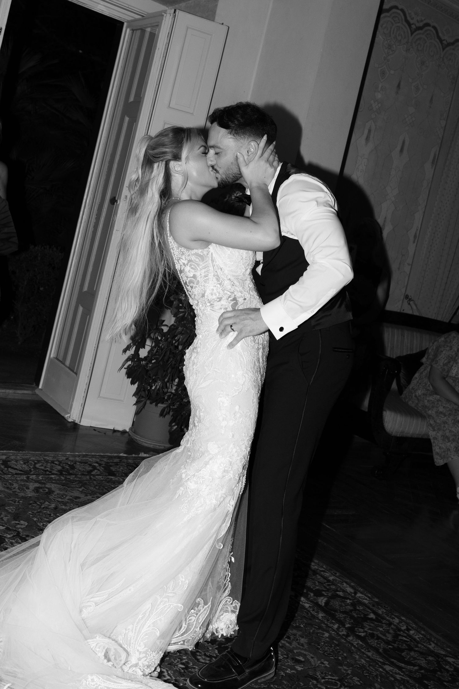 A black and white image showing a bride and groom embracing and kissing at their wedding reception.