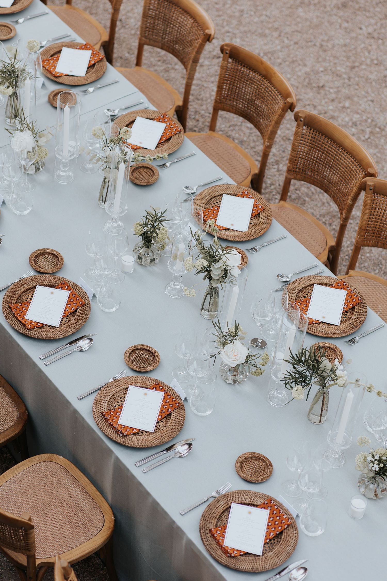 A table setting with woven rattan chairs, white linens, and floral centerpieces for a rustic, outdoor wedding reception.