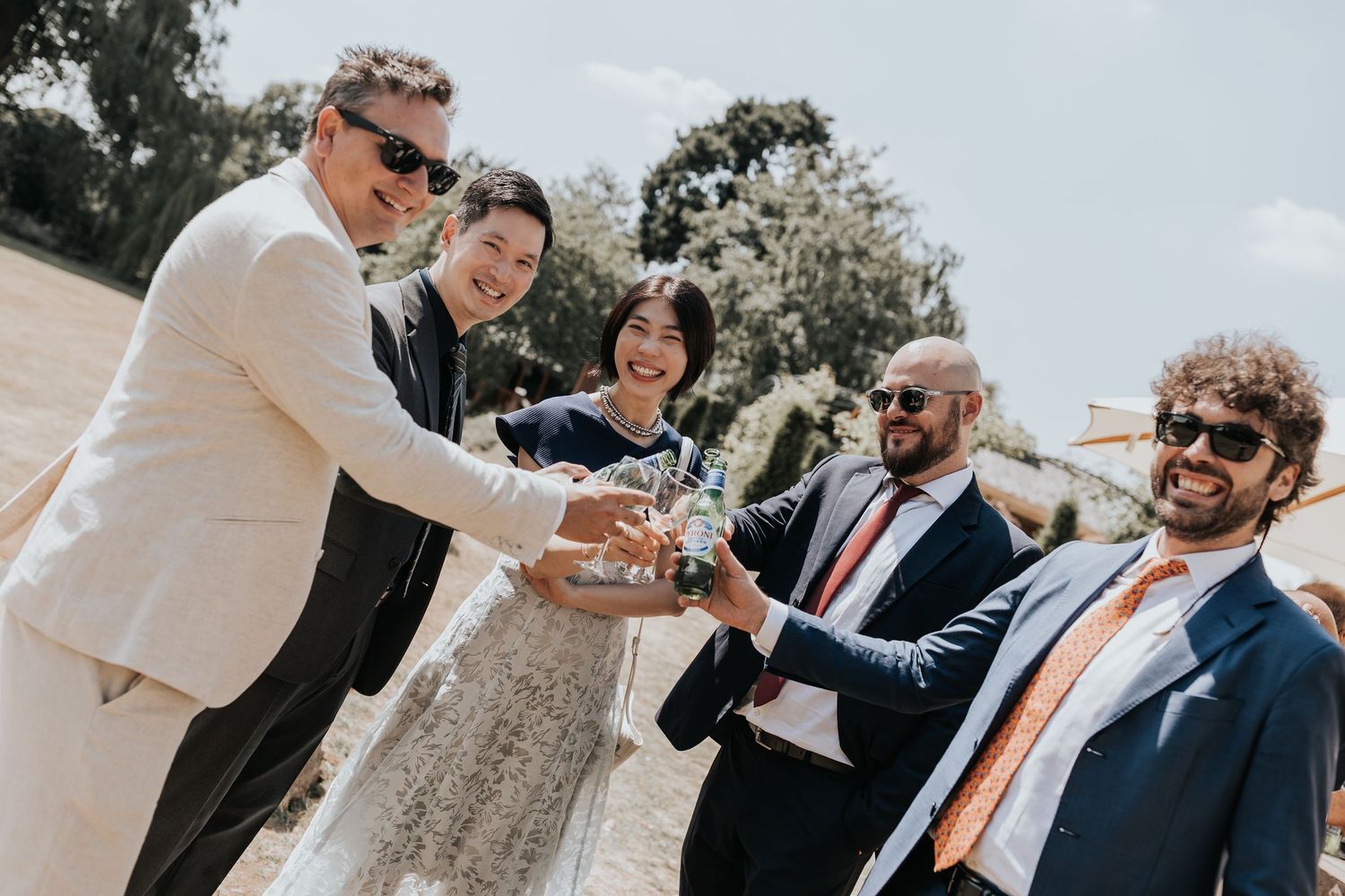 Wedding guests clinking glasses outdoors, celebrating with the bride and groom on a sunny day.