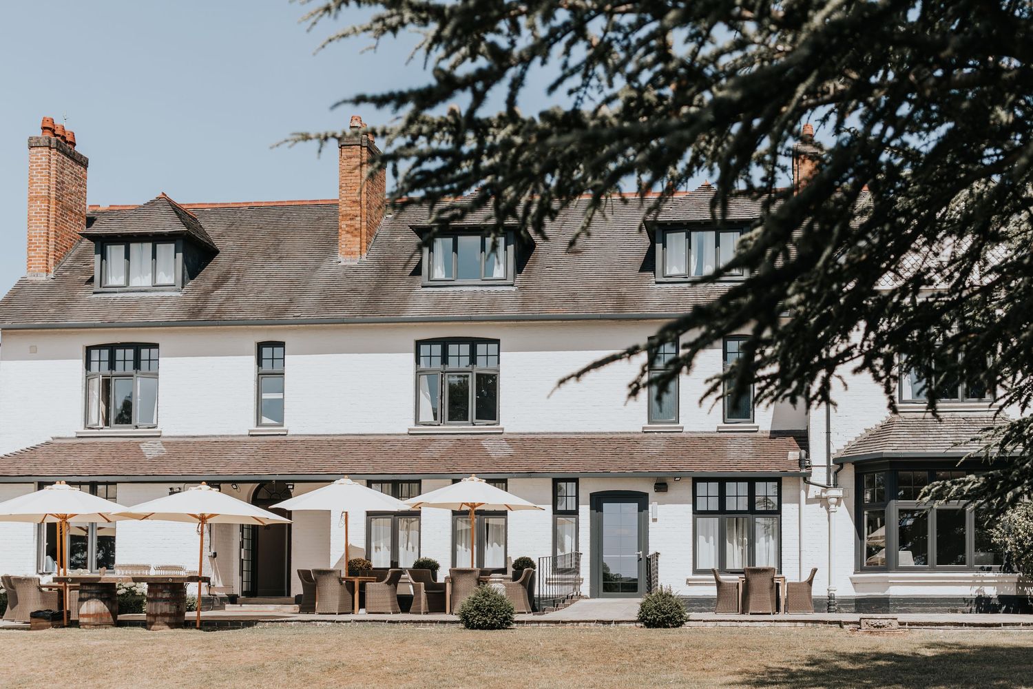White country house hotel with outdoor seating, parasols, and manicured lawns framed by cedar trees on a sunny day.