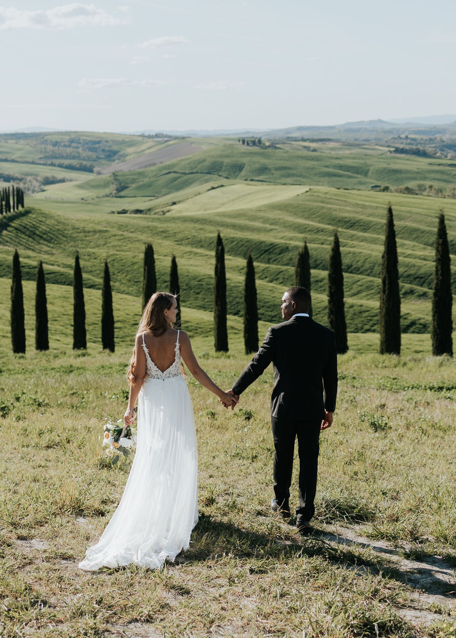 Bride and groom holding hands overlooking rolling Tuscan hills with cypress trees on their wedding day.