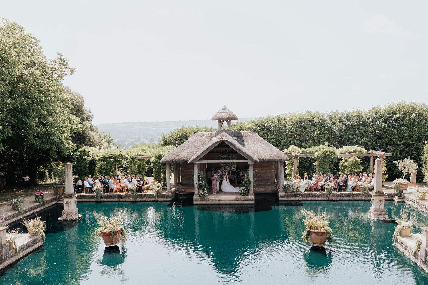 Elegant outdoor wedding ceremony by a reflecting pool with a gazebo, lush greenery, and guests seated in a garden venue.