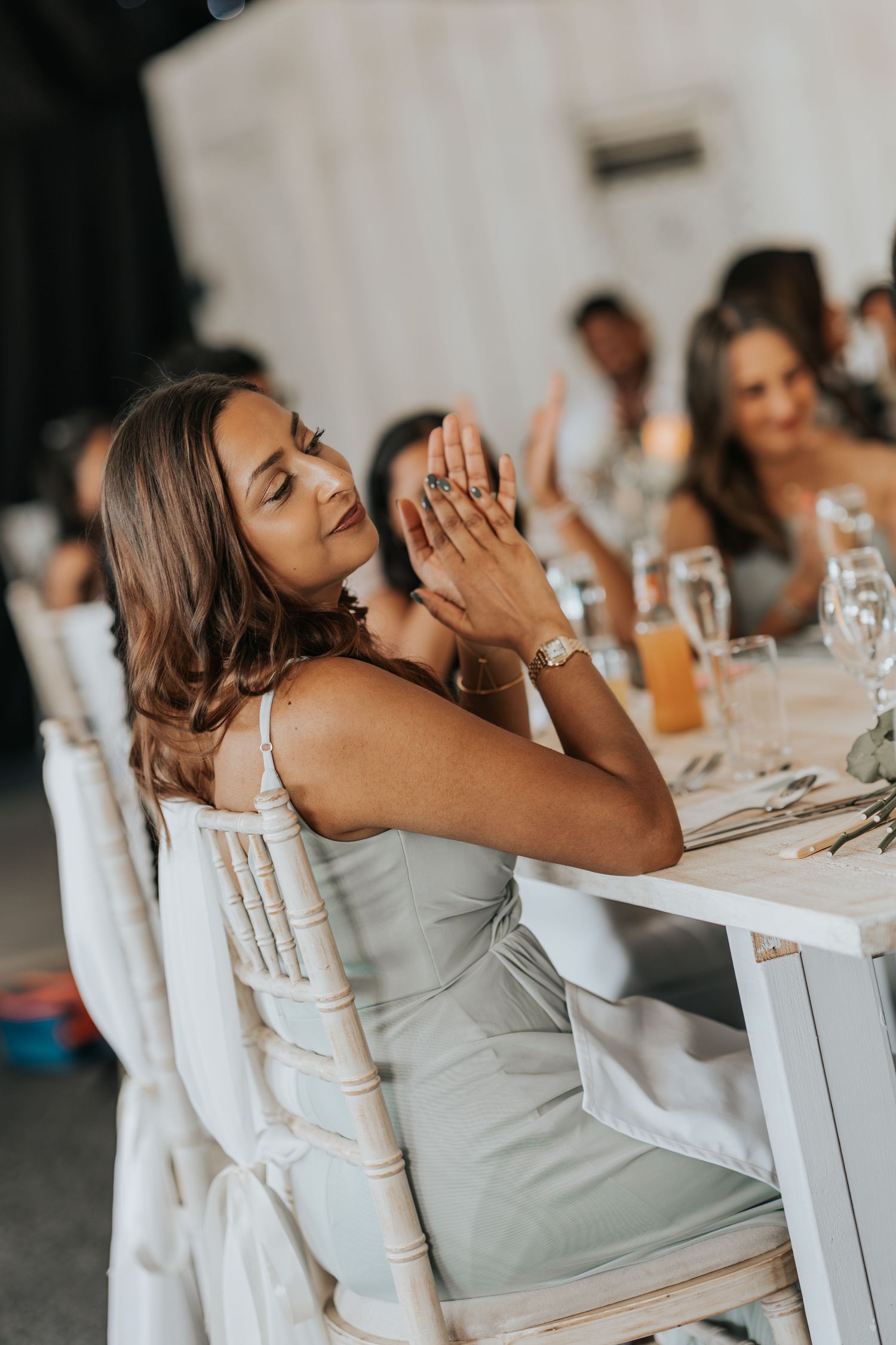 Woman in sage green dress clapping at elegant wedding reception dinner table with guests in background.
