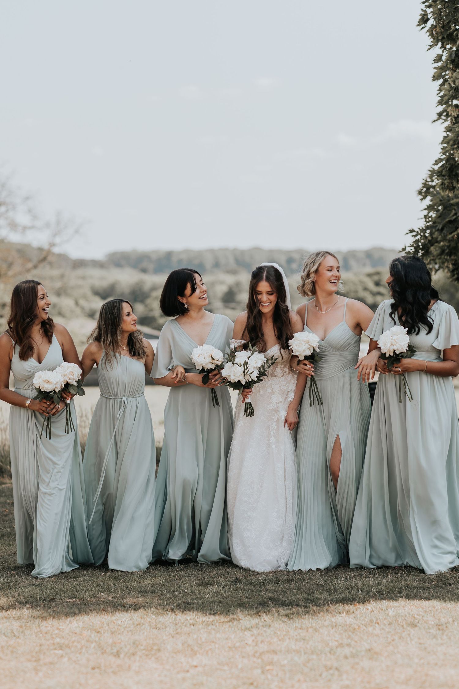 Bride in white lace gown with five bridesmaids in sage green dresses holding white bouquets outdoors.