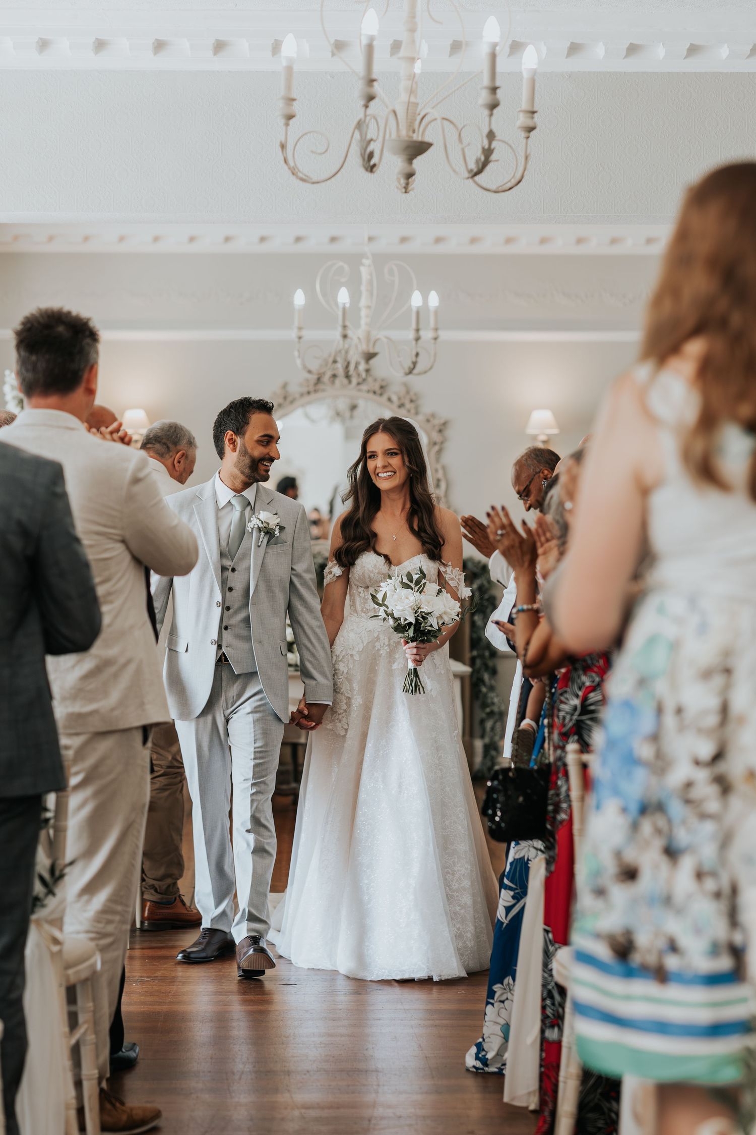 A bride and groom walk down the aisle surrounded by wedding guests in a formal indoor ceremony.