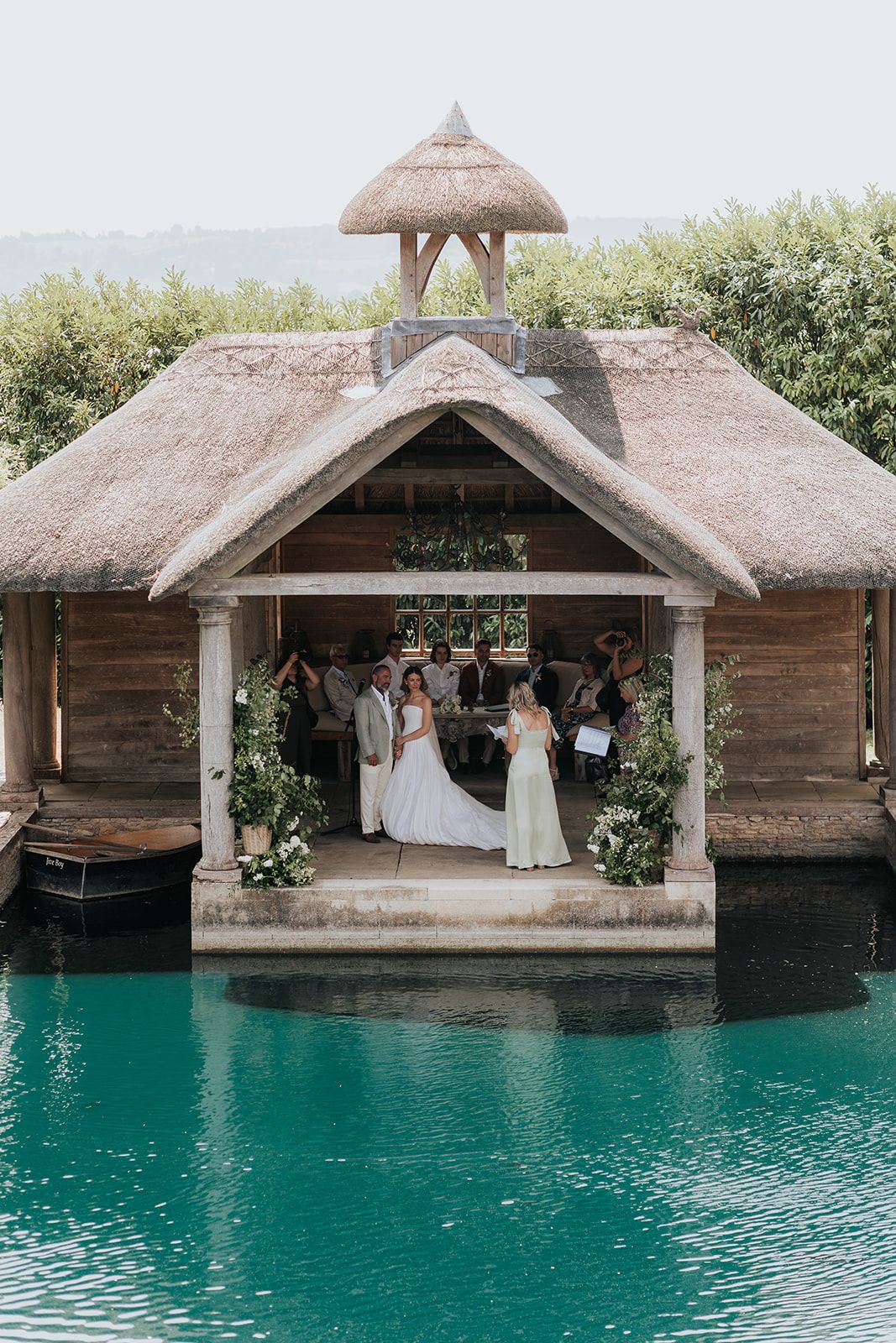 Wedding ceremony in a thatched boathouse over turquoise water, bride and guests gathered among floral decorations.