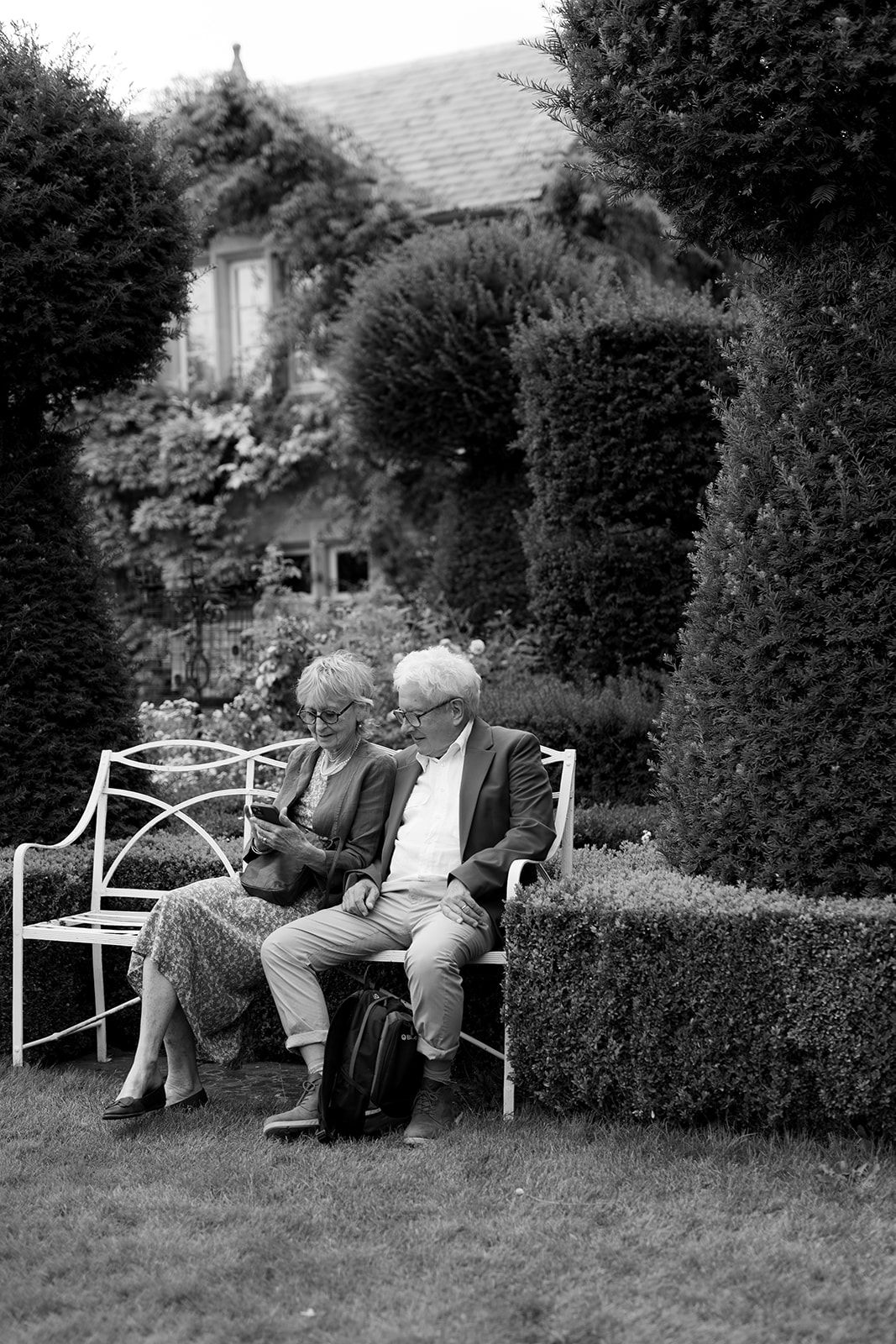 Elderly couple sitting together on a white garden bench surrounded by lush hedges and ivy-covered house in black and white.