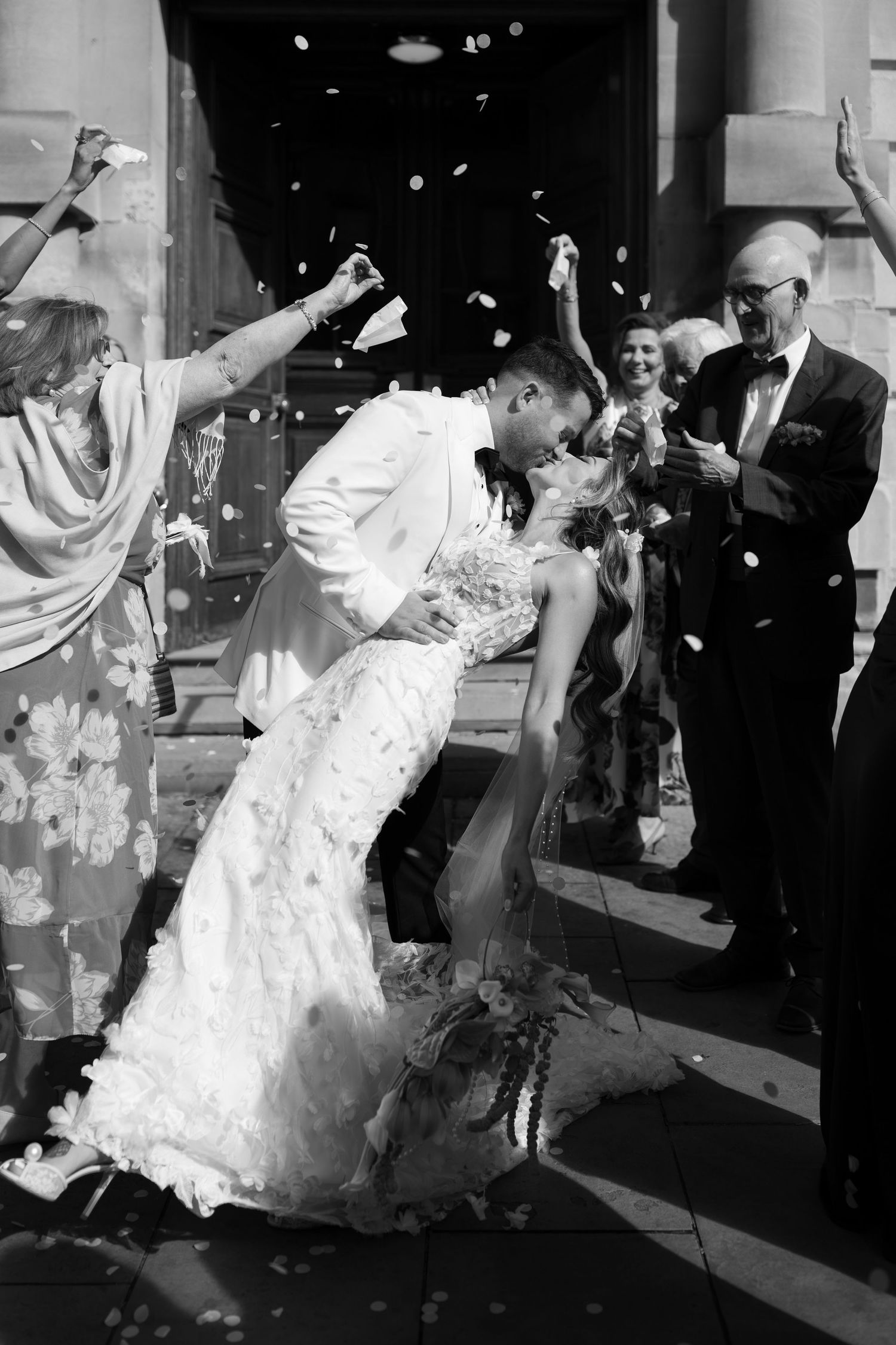 Black and white photo of newlyweds sharing a dip kiss while guests toss confetti outside a church.