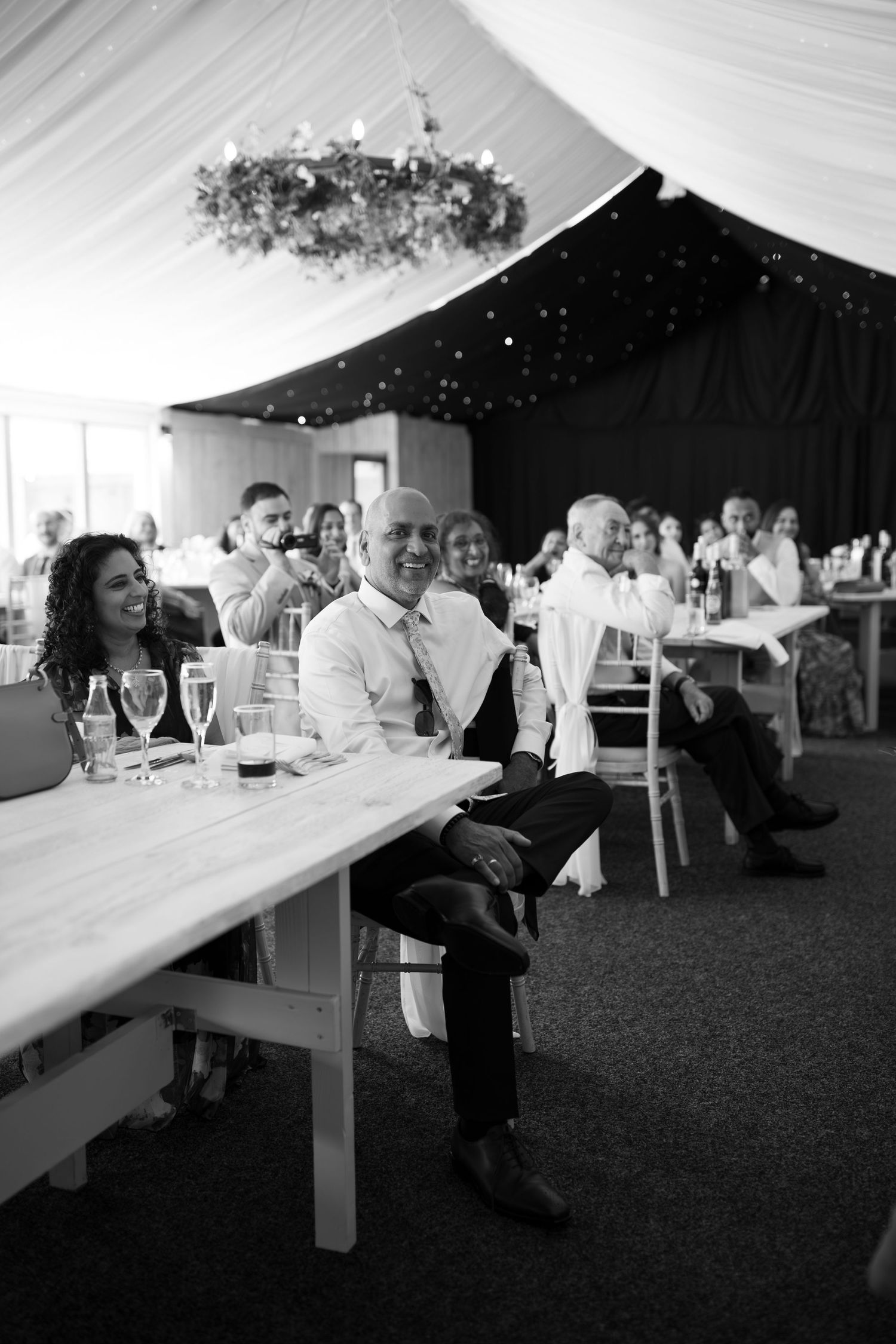 Black and white photo of wedding guests laughing and enjoying speeches at elegant marquee reception venue.