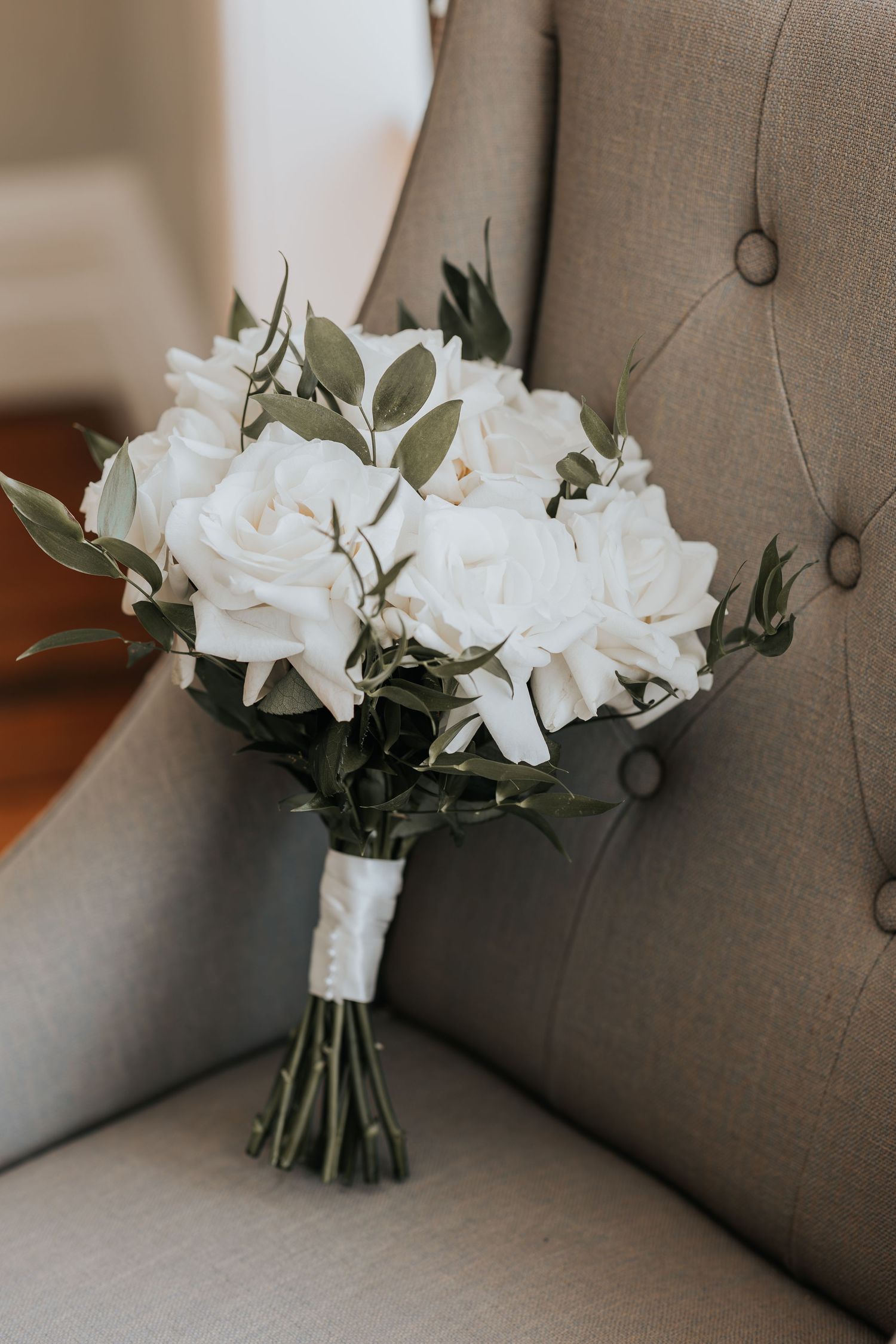 Elegant bridal bouquet of white roses and greenery wrapped in white ribbon, resting on a tufted gray chair.