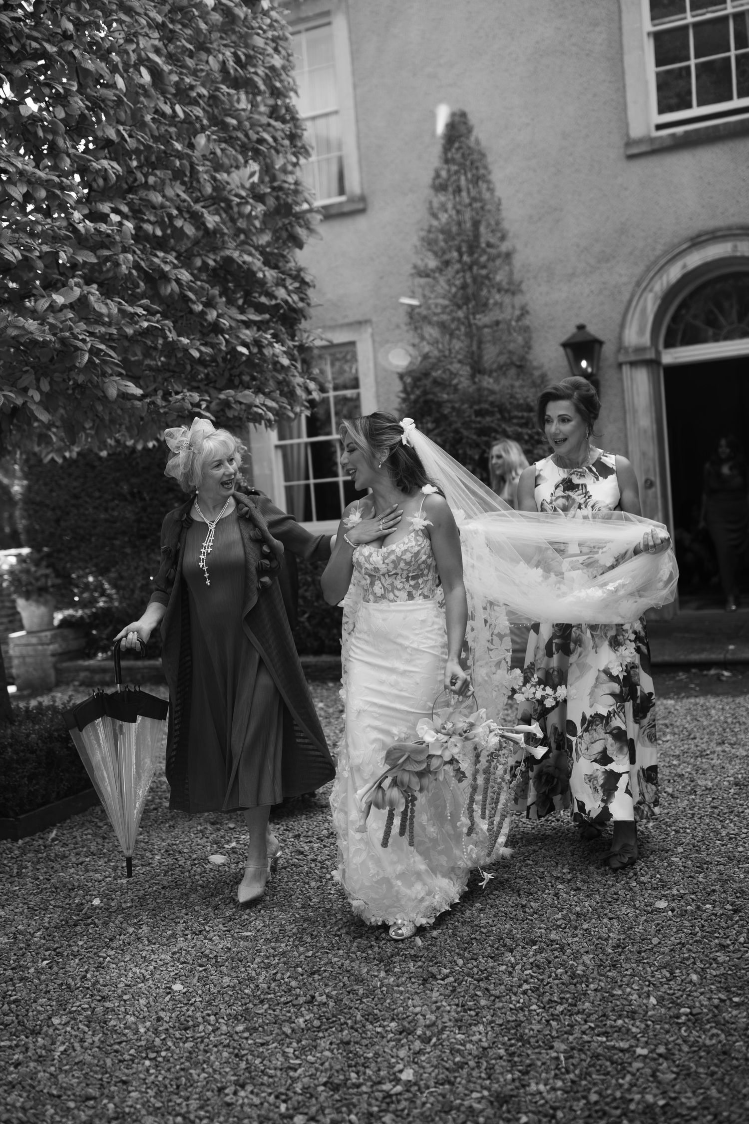 Black and white photo of a bride in a lace gown with veil being held by attendants outside an elegant manor.