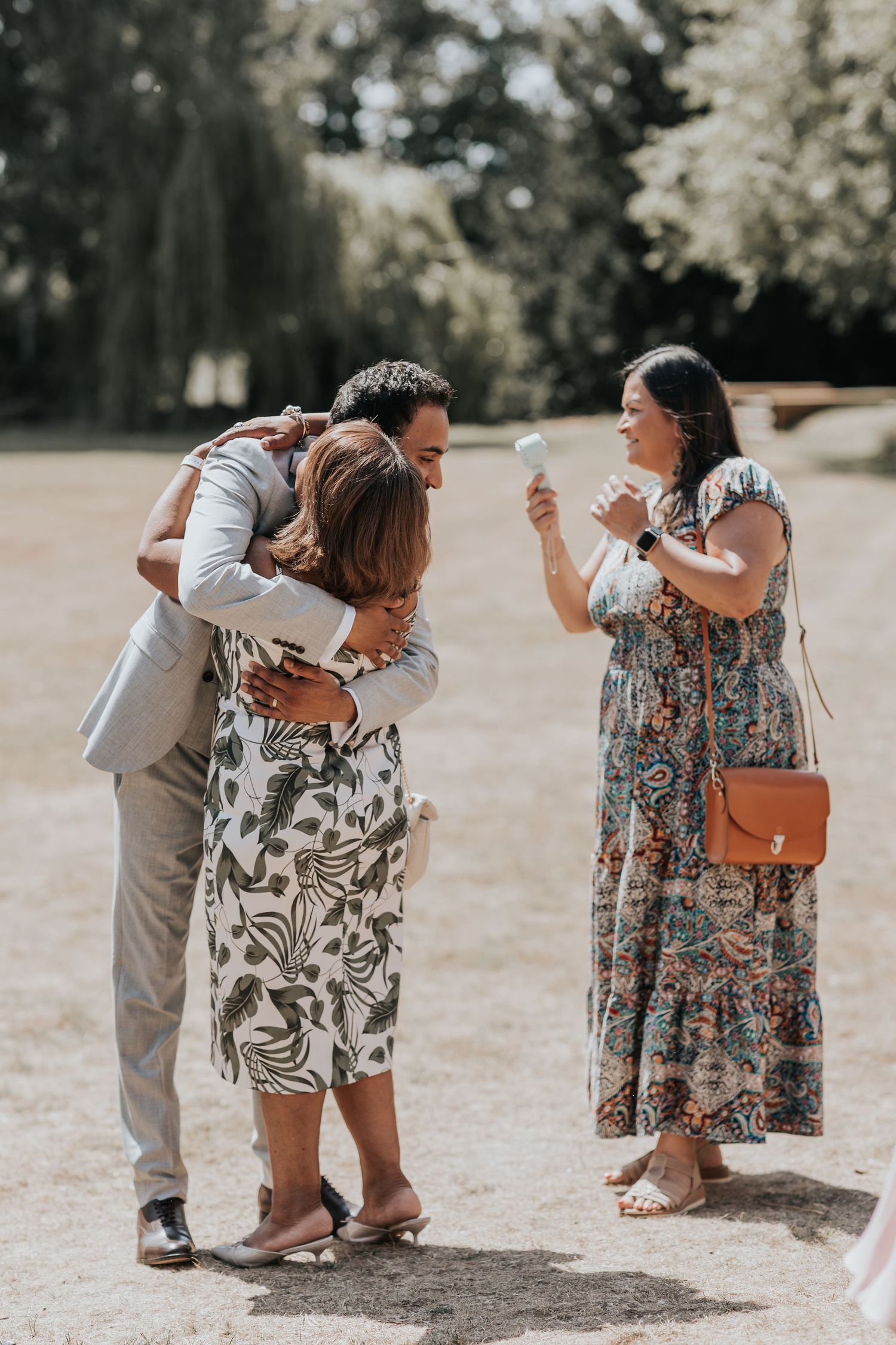 Man in suit hugging woman in floral dress outdoors while another woman in maxi dress photographs them at a wedding.