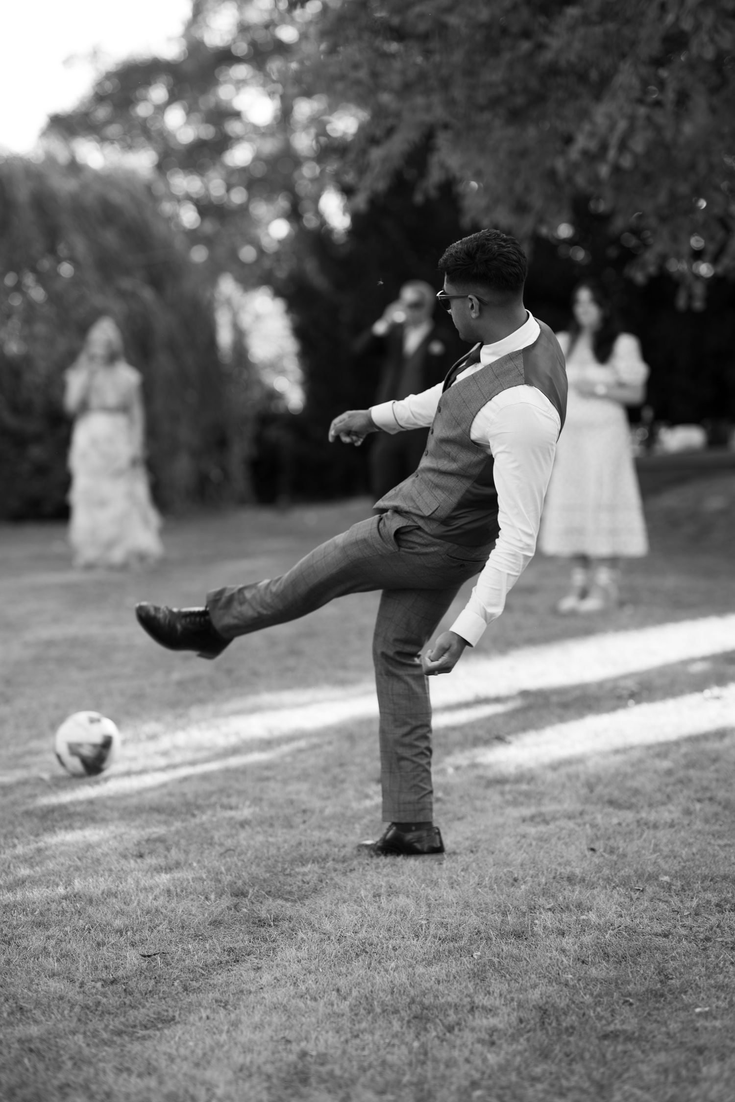 Man in formal vest kicks a soccer ball on grass at an outdoor wedding reception in black and white photo.