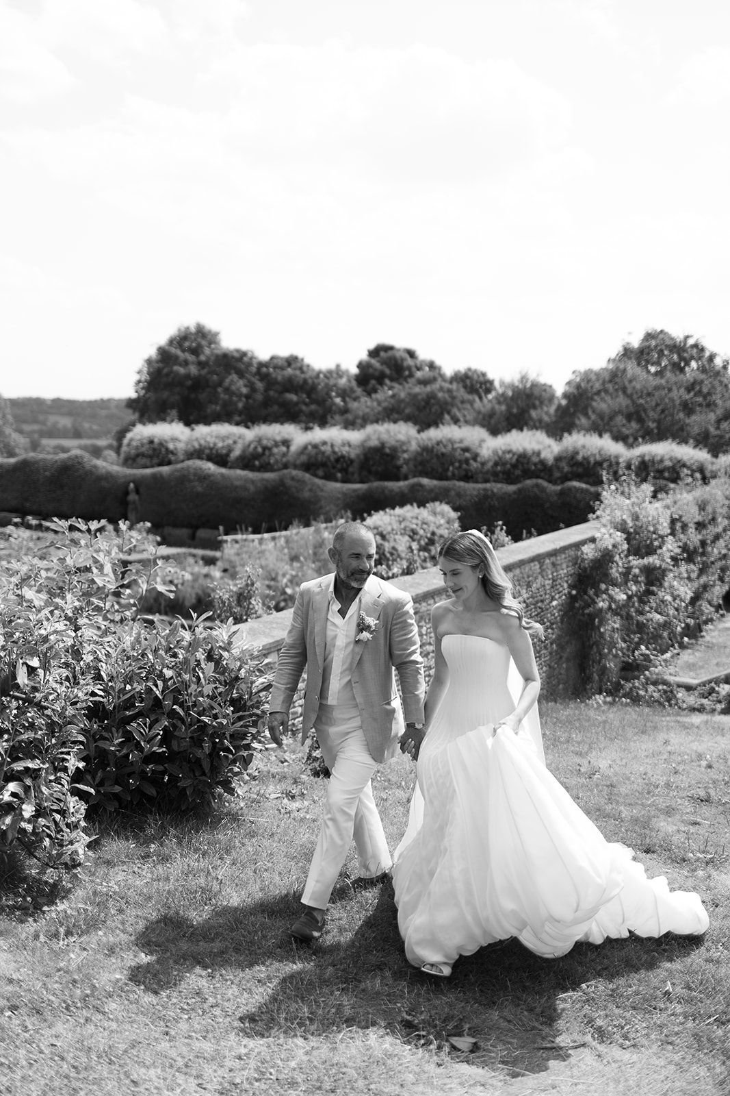 Black and white photo of a bride and groom holding hands walking through a lush garden on their wedding day.