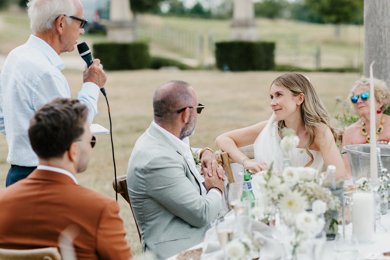 Guests smile and listen during an outdoor wedding reception speech at an elegant garden venue.