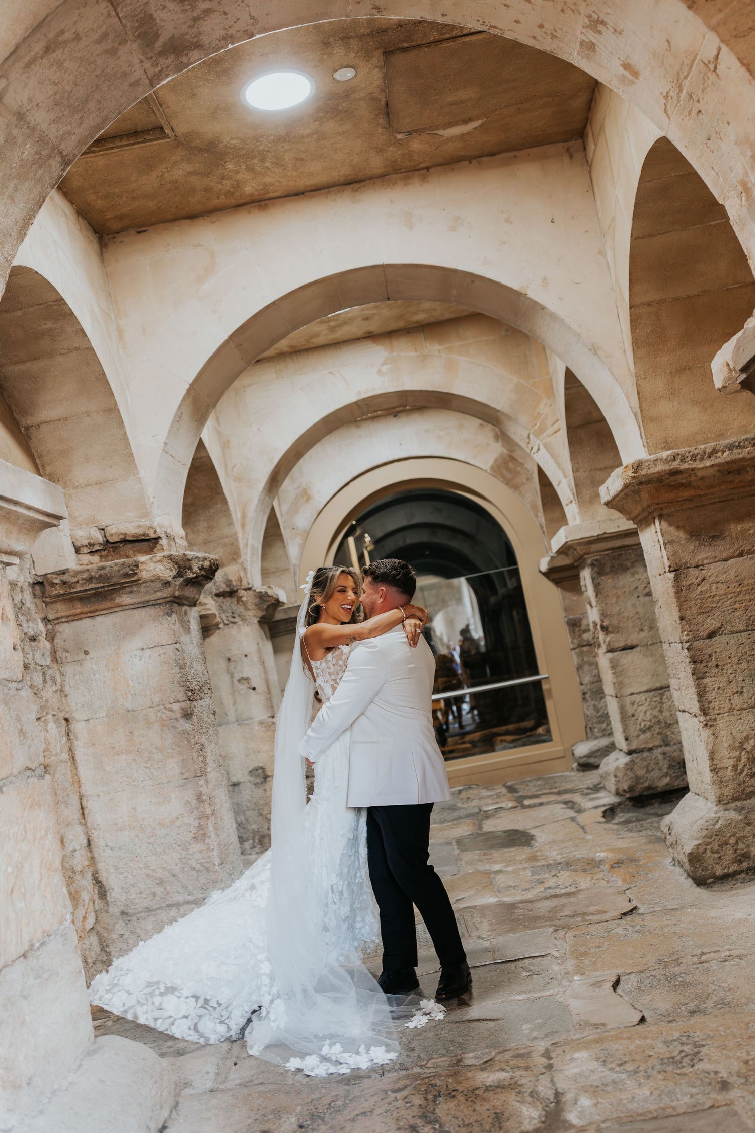 Bride and groom embrace under stone arches in a historic venue, bride wearing a lace train gown.