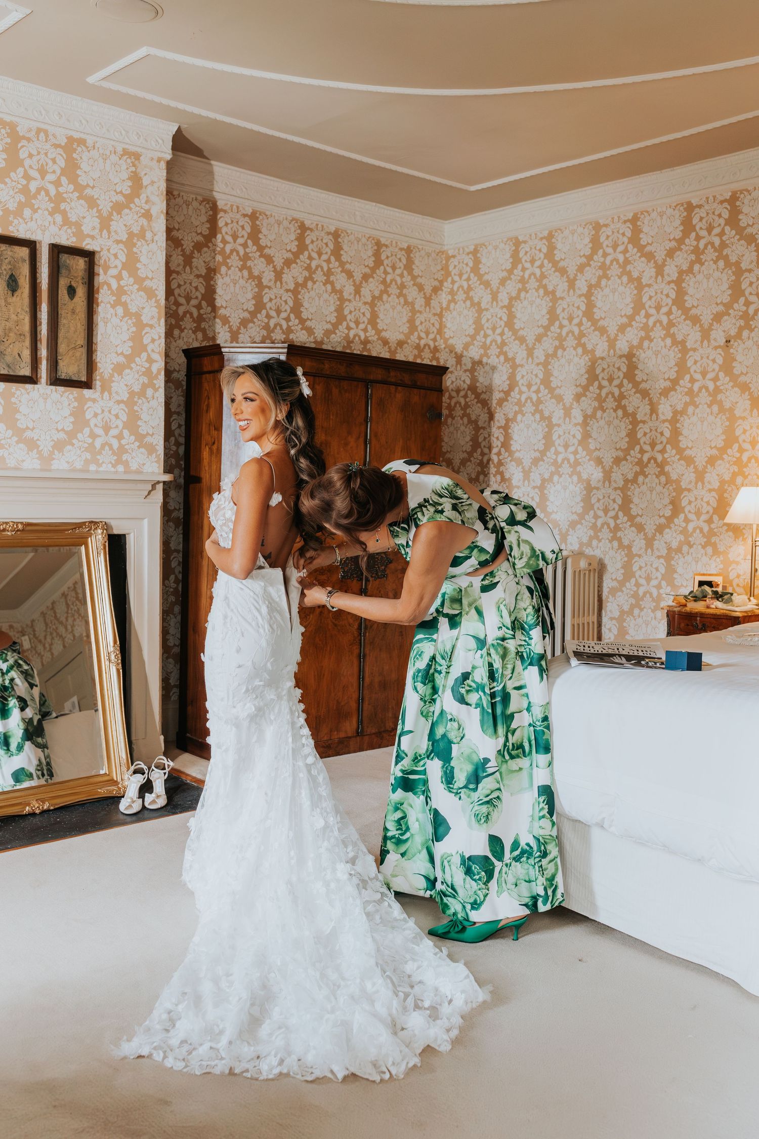 Bride in lace mermaid wedding dress being helped by woman in green floral dress in elegant wallpapered room.