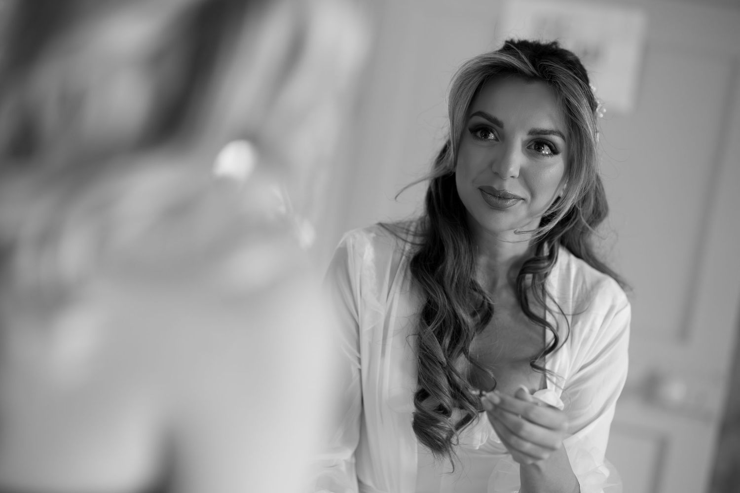 Black and white portrait of a bride in a white robe, gazing into a mirror while getting ready on her wedding day.