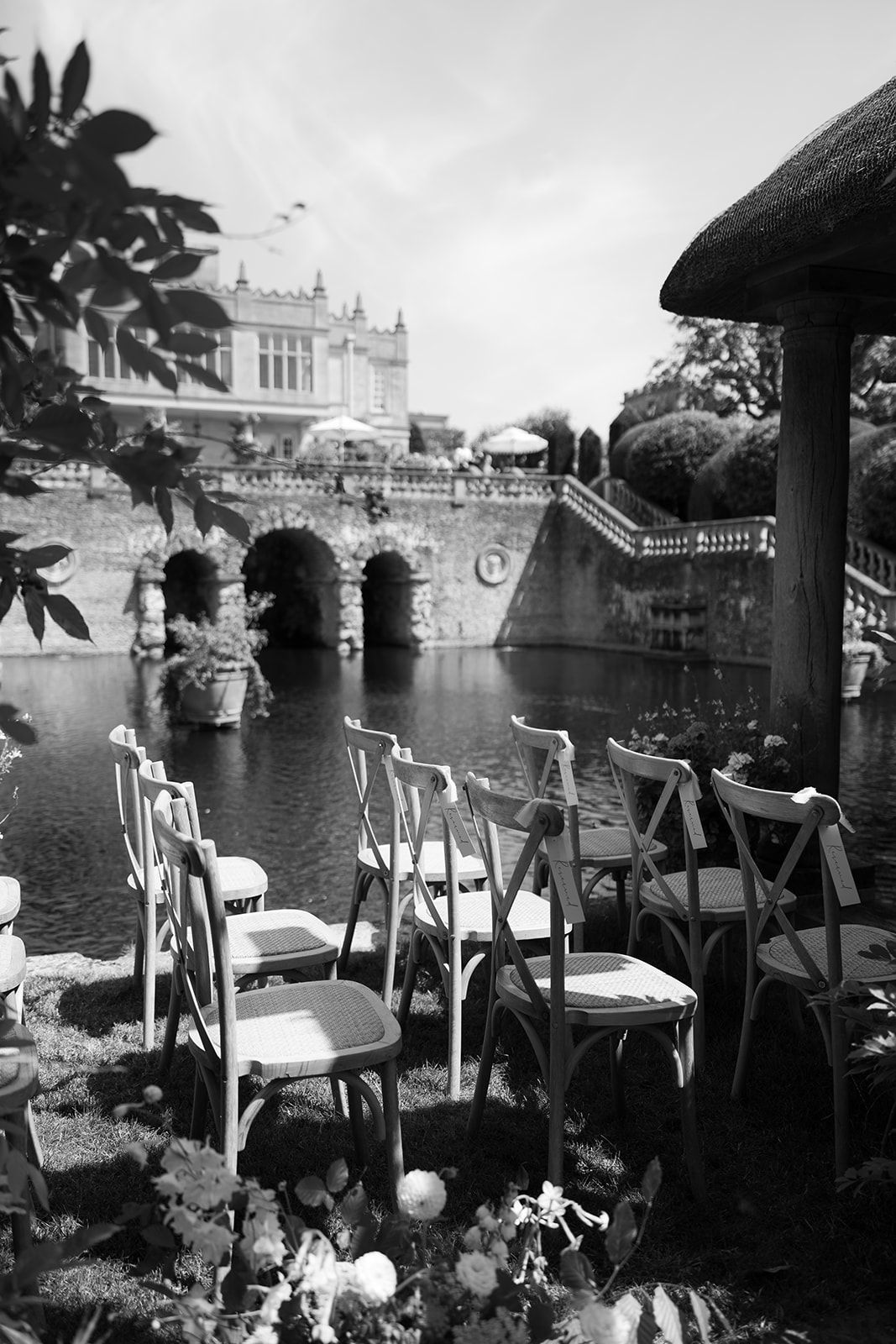 Black and white photo of elegant chairs arranged by a ornate stone castle pond for an outdoor wedding ceremony.