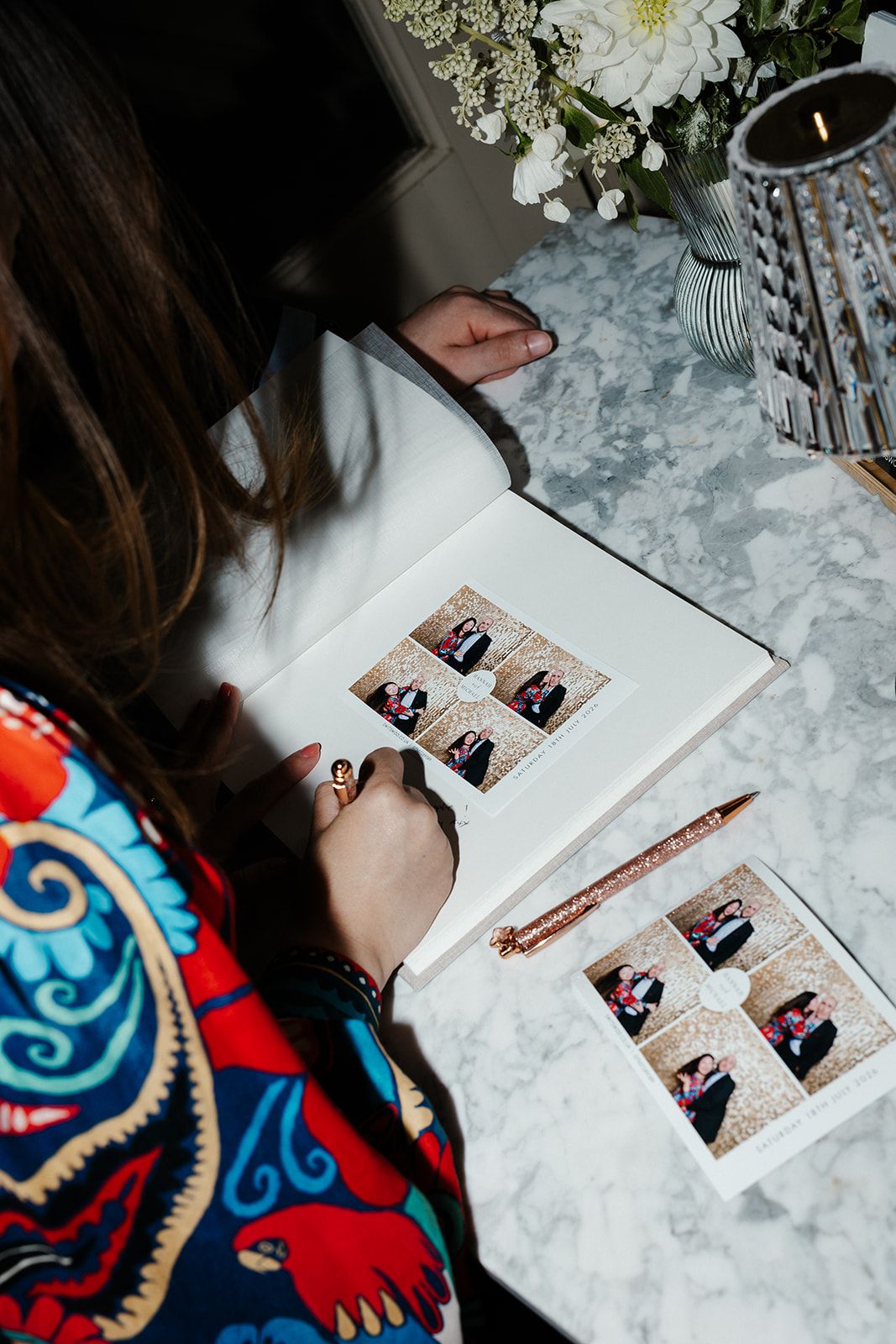 Woman in colorful dress reviews photo booth pictures in a wedding guest book on a marble table with flowers.