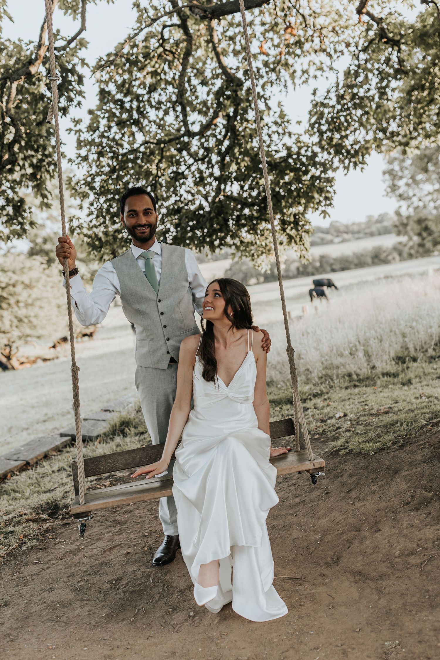 Bride in white gown sits on tree swing as groom in gray suit stands behind her at outdoor wedding.
