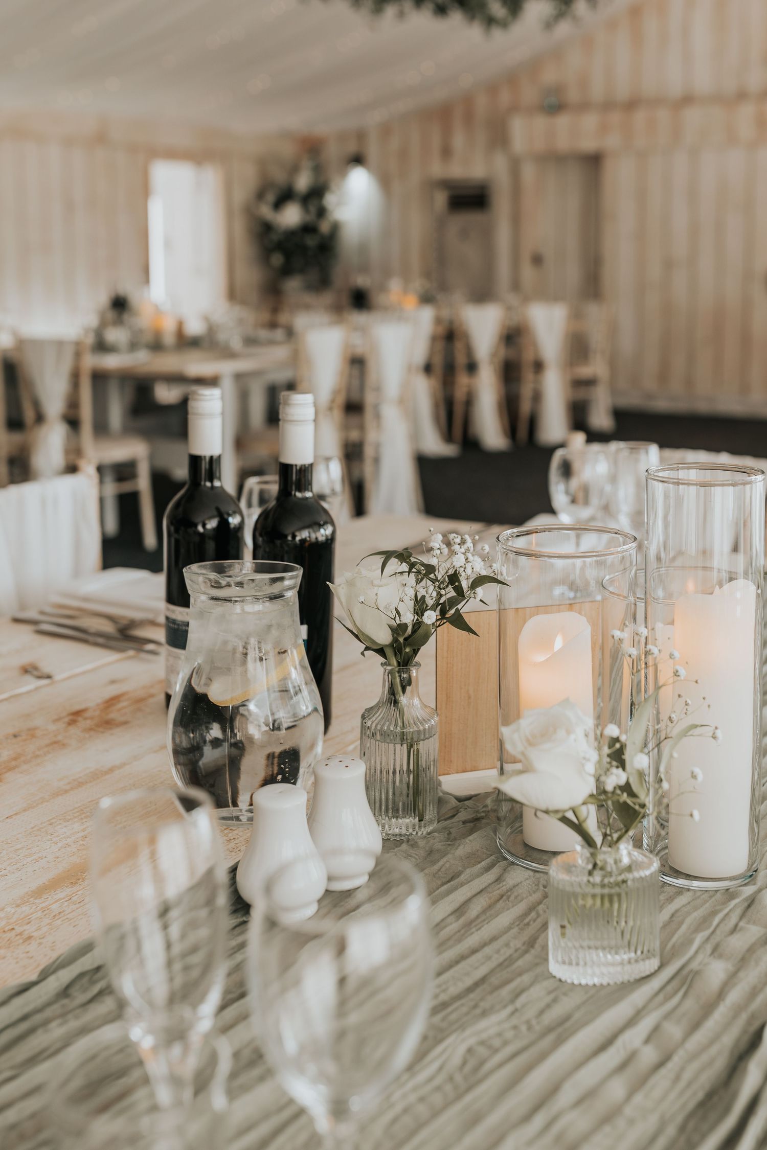 Elegant wedding reception table with white candles, glass vases, wildflowers, and wine bottles on marble surface.