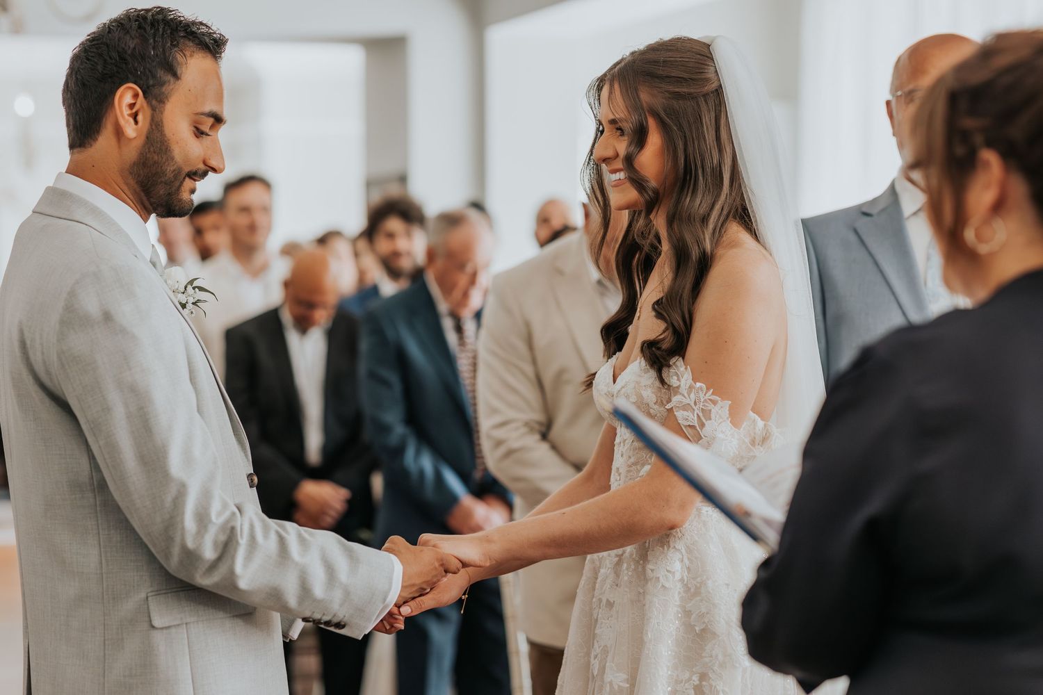Bride and groom holding hands and smiling during outdoor wedding ceremony with guests in background.