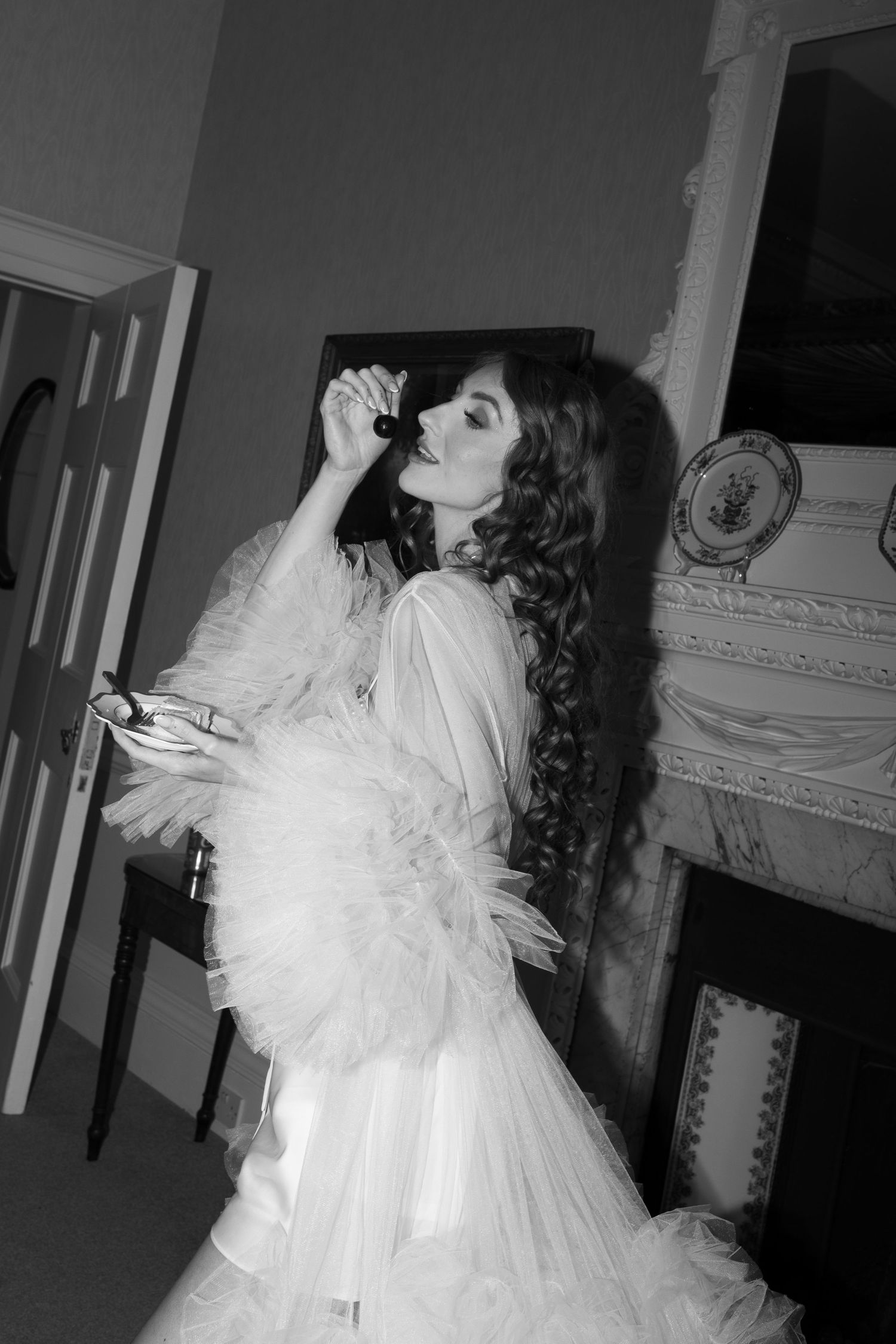 A woman in a white, feathered dress stands in a room, posing with her hands in her hair.