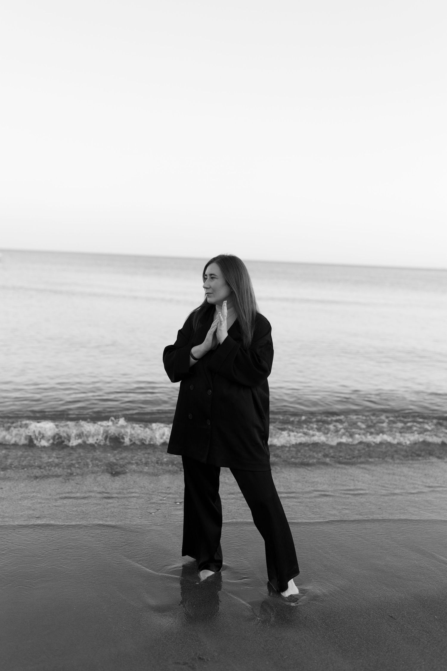 A woman in a black outfit standing in the water on a beach, with the ocean in the background.