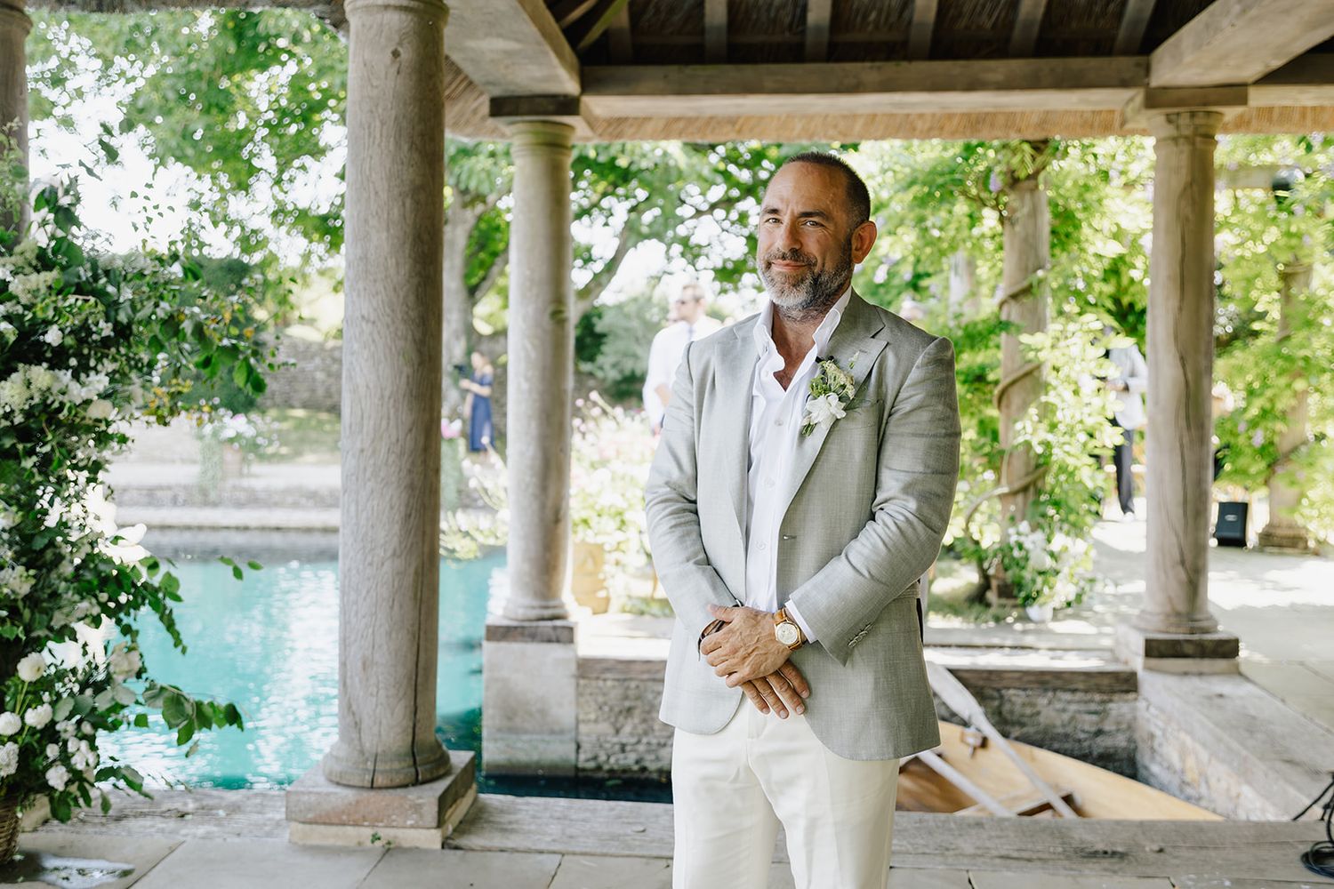 Groom in light gray suit with white boutonniere stands under stone pavilion near pool at outdoor wedding venue.