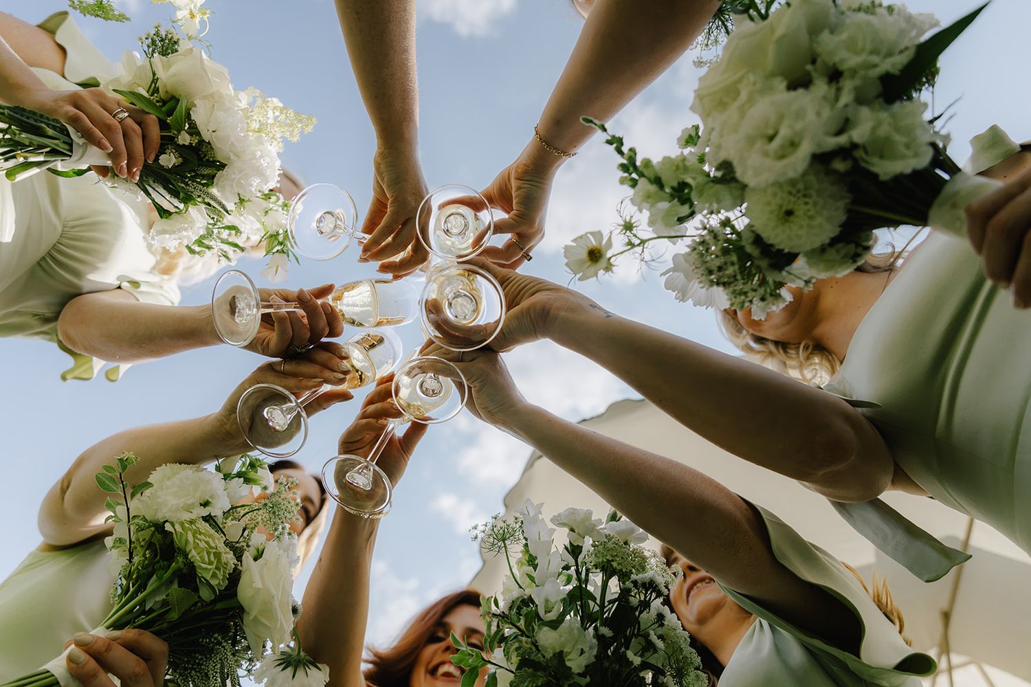 Bridesmaids in sage green dresses toasting champagne glasses with white floral bouquets against a blue sky.
