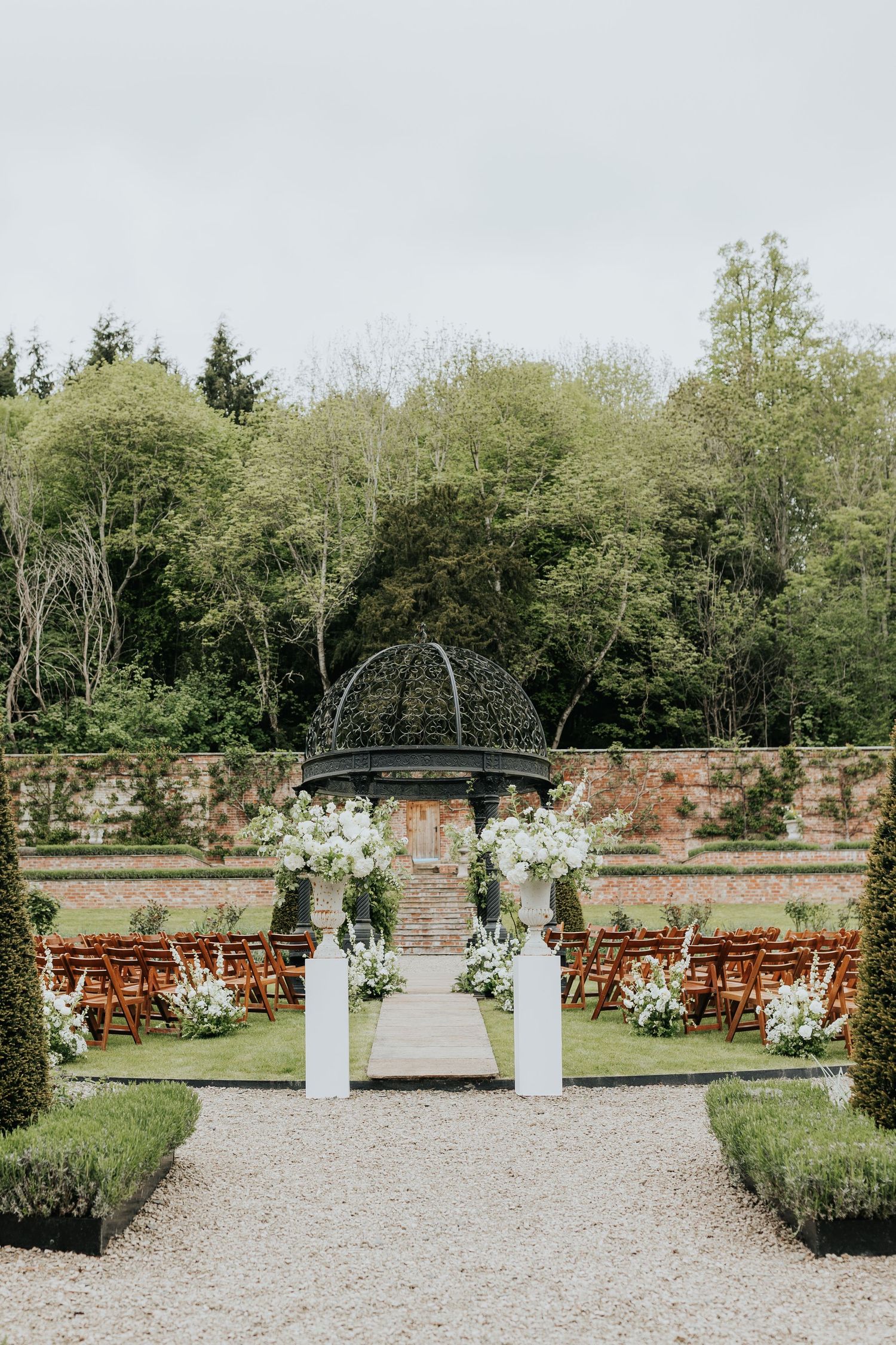 Elegant outdoor wedding ceremony setup in a walled garden with white floral arrangements, wooden chairs, and a domed gazebo backdrop.