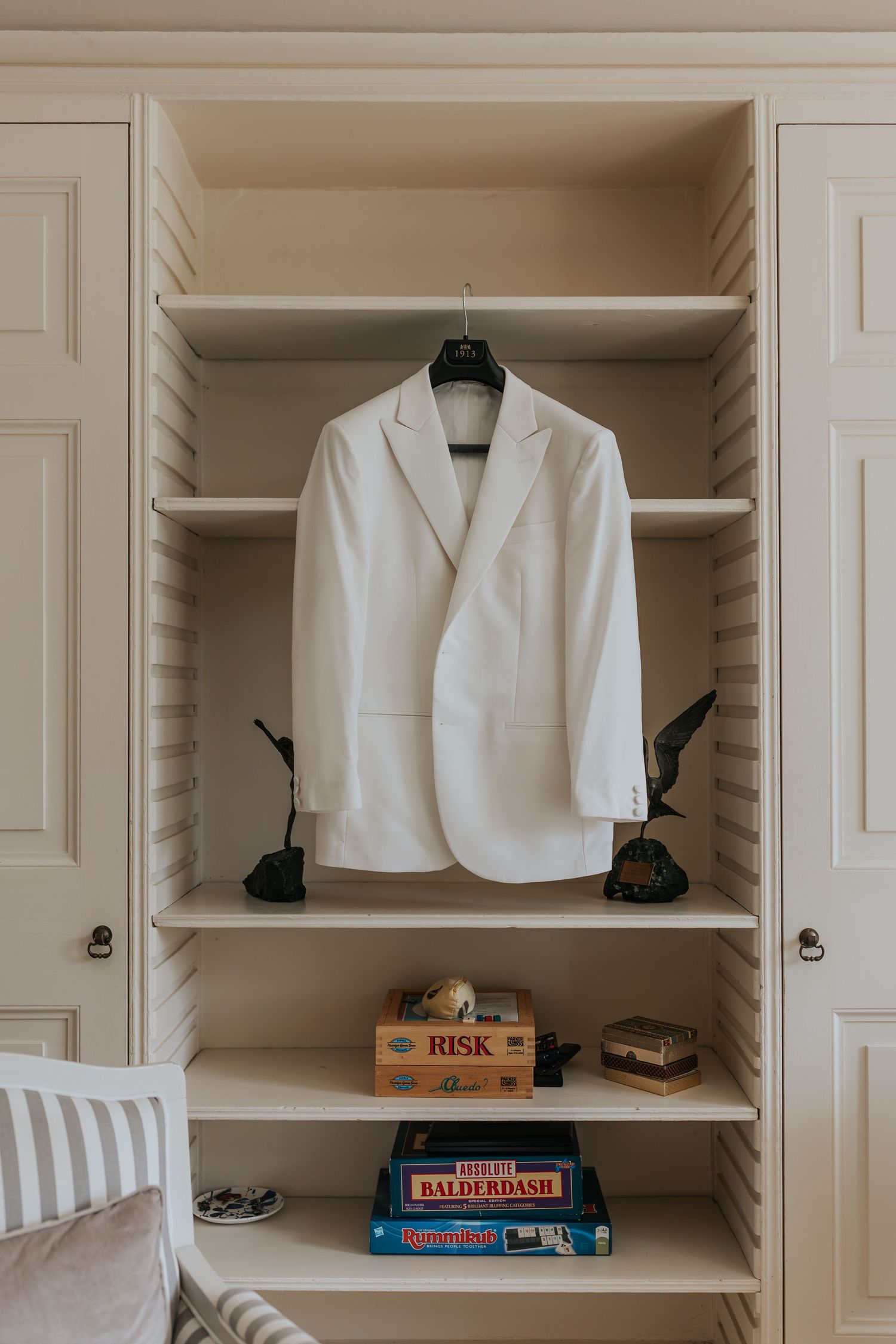 White blazer hanging in a cream built-in wardrobe with board games including Risk and Balderdash on lower shelves.