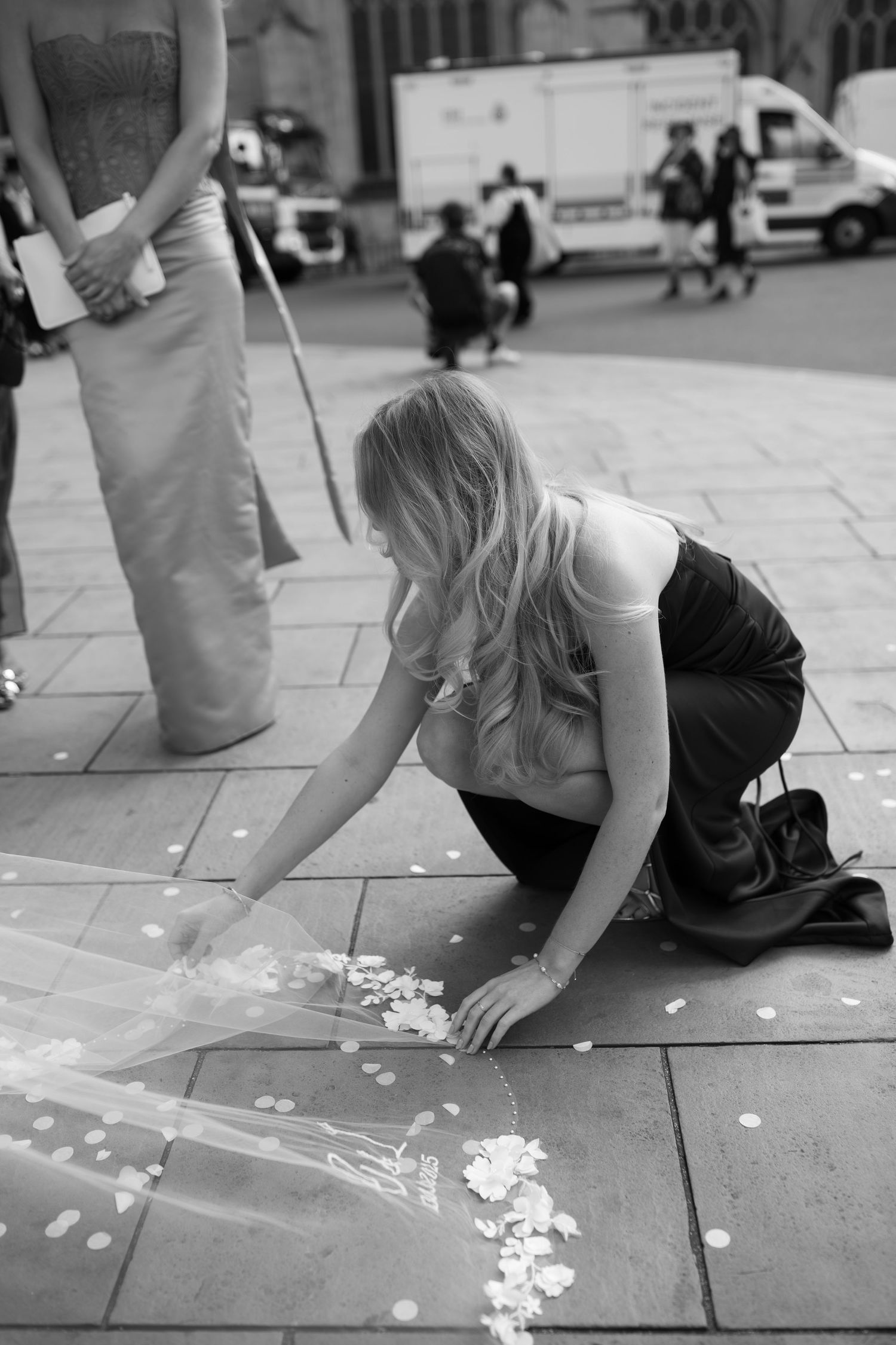 Black and white photo of a woman kneeling on pavement arranging a bridal veil and flower petals outside a church.