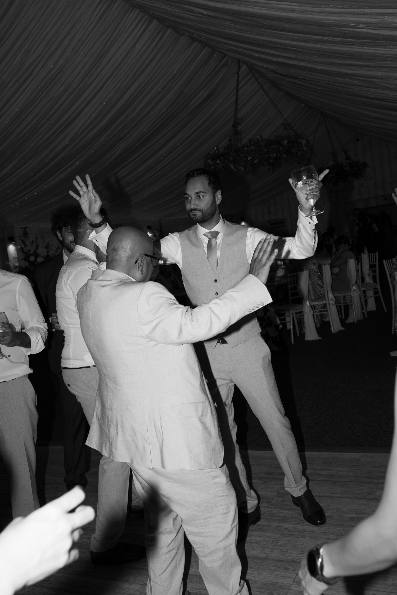Black and white photo of men in suits dancing at a wedding reception in a decorated marquee tent.