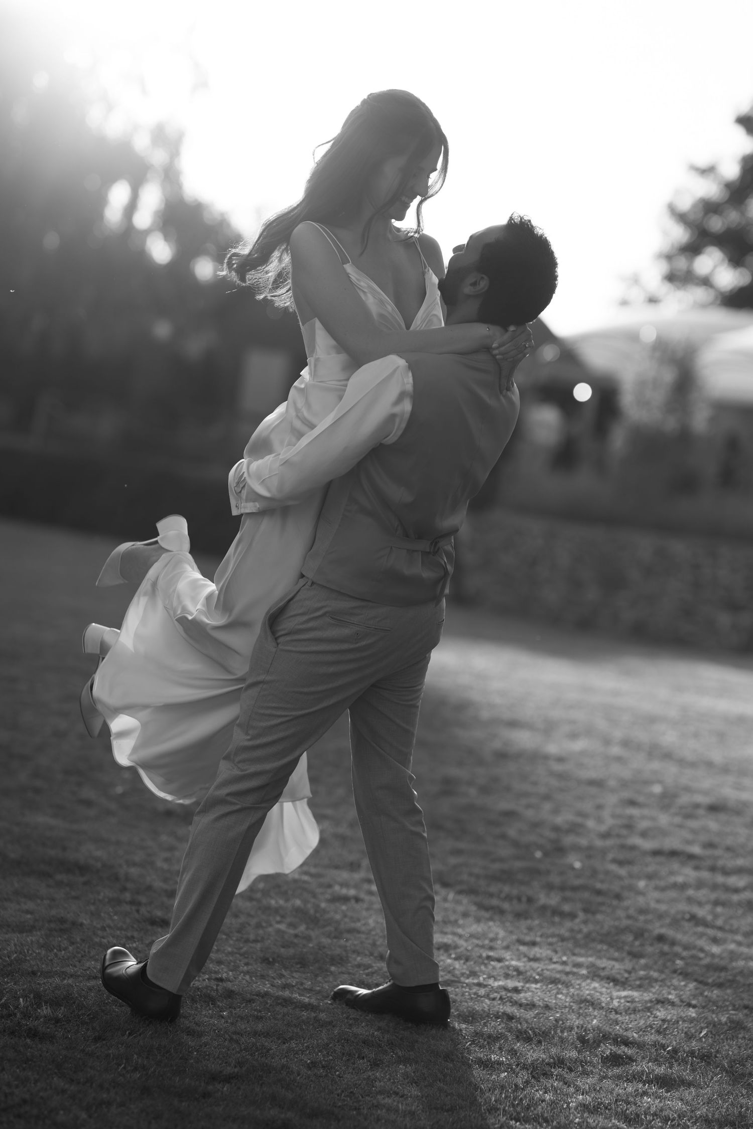 Black and white photo of a groom lifting his bride in a flowing dress outdoors at sunset.