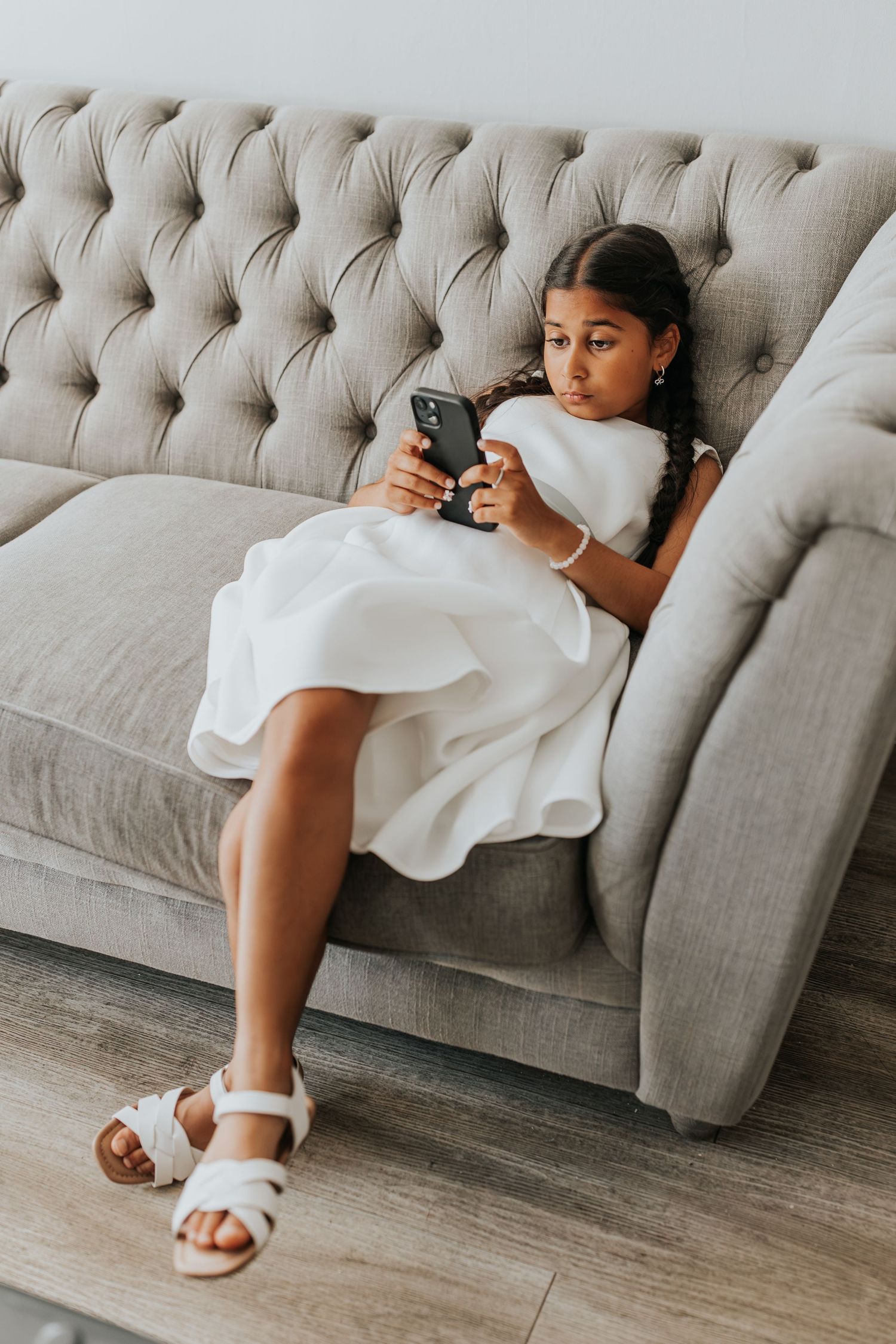 Young girl in white dress and sandals relaxing on a tufted gray sofa while looking at her smartphone.