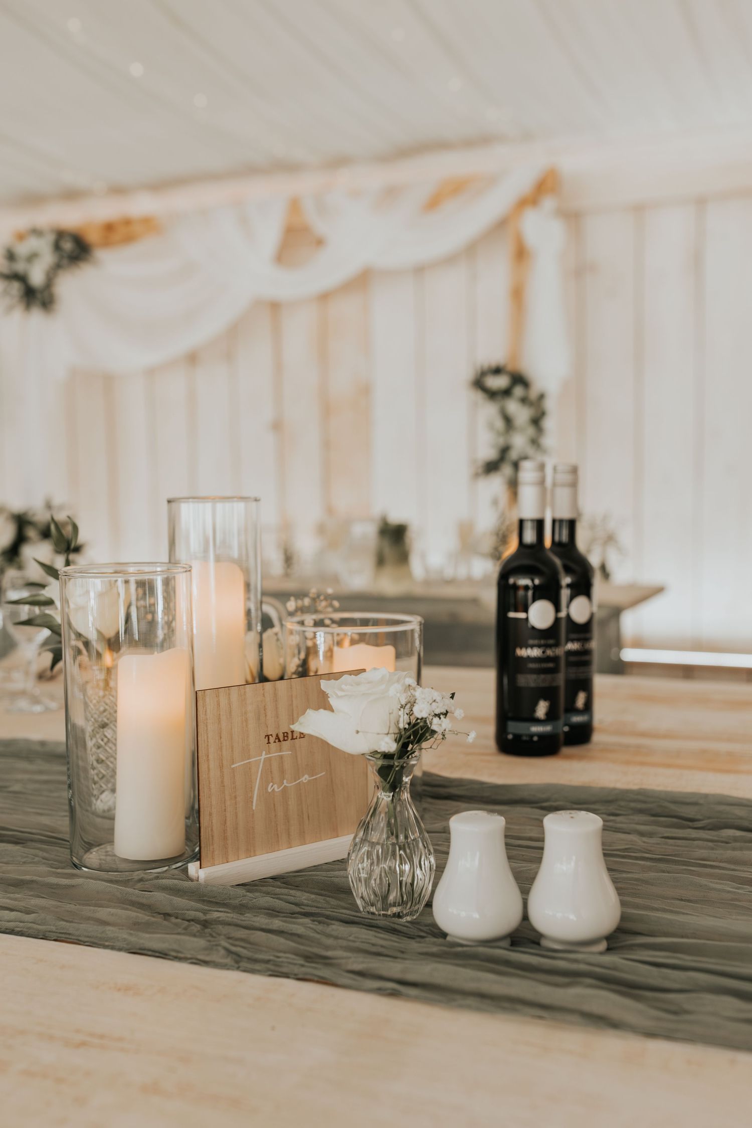 Elegant wedding reception table with white candles, floral vase, menu card, salt and pepper shakers, and wine bottle.
