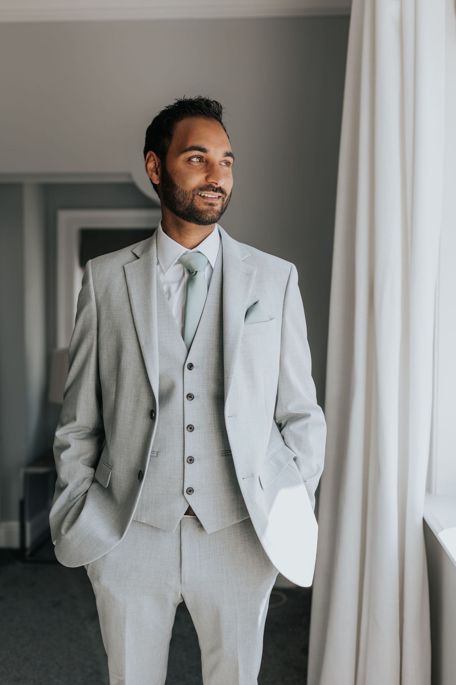Groom in light gray three-piece suit with mint tie and pocket square standing by window.